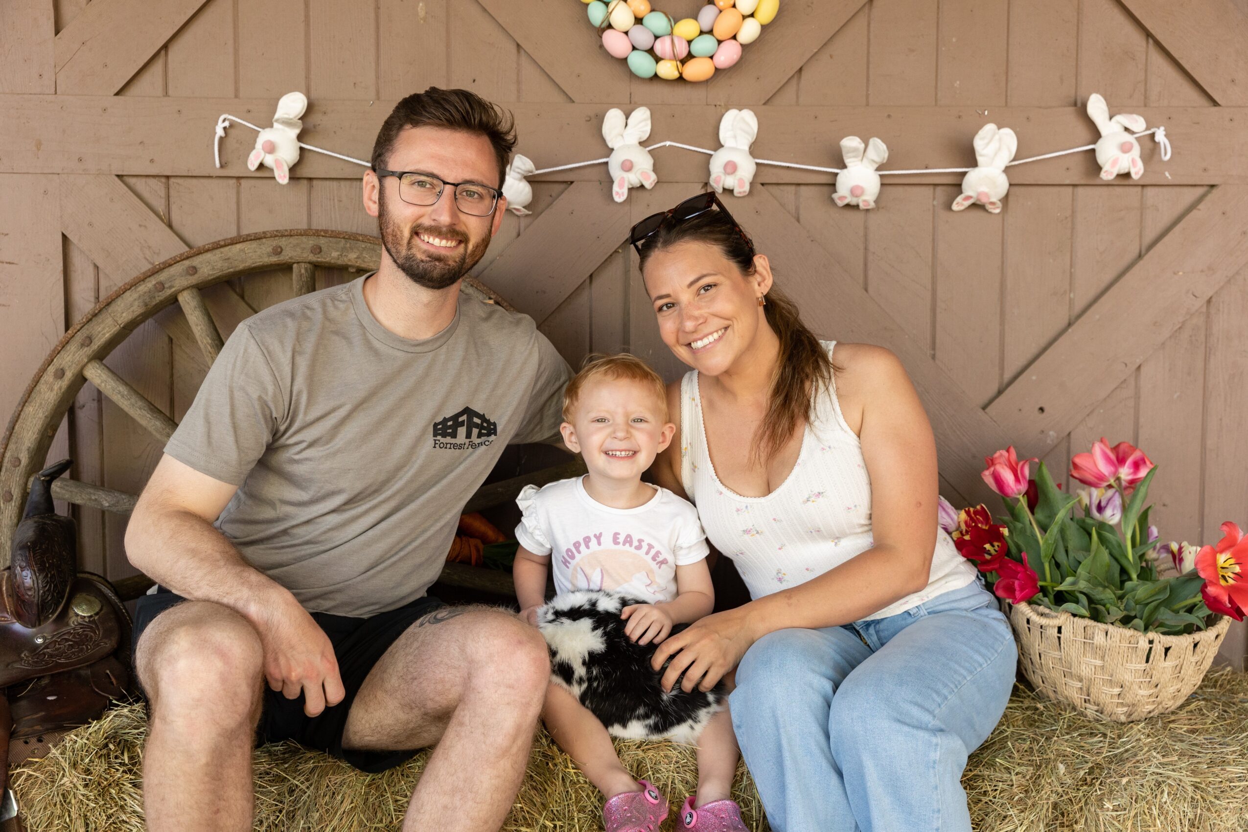 Family of three sitting on hay, smiling, with decorated background and flowers, celebrating Easter or spring, indoors.