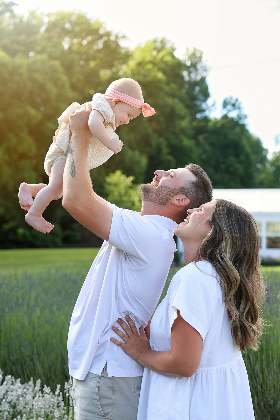 Man and woman outdoors with a baby, man lifting baby, woman smiling, green trees in background