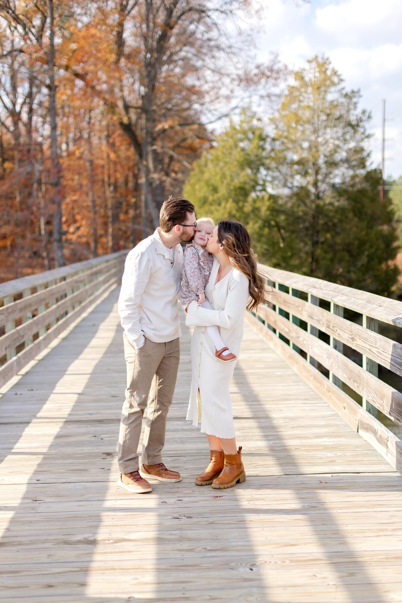 A couple stands on a wooden bridge, kissing, with trees in autumn colors in the background.