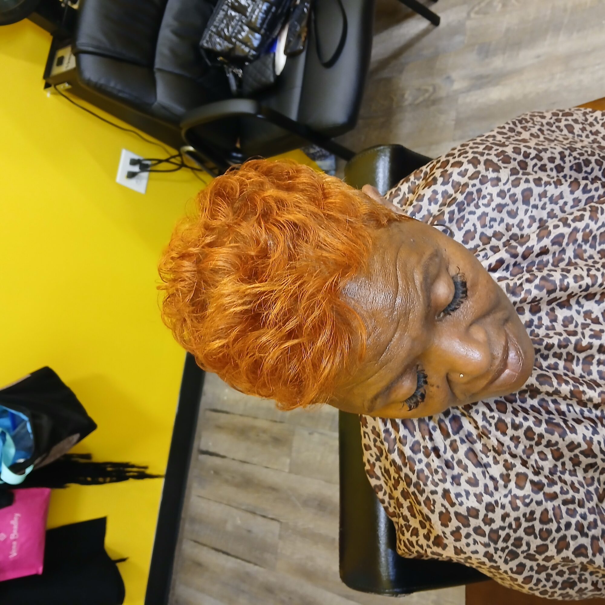 Woman with short, curly, orange hair sitting in a salon chair, wearing a leopard print cape, with a yellow wall in the background.
