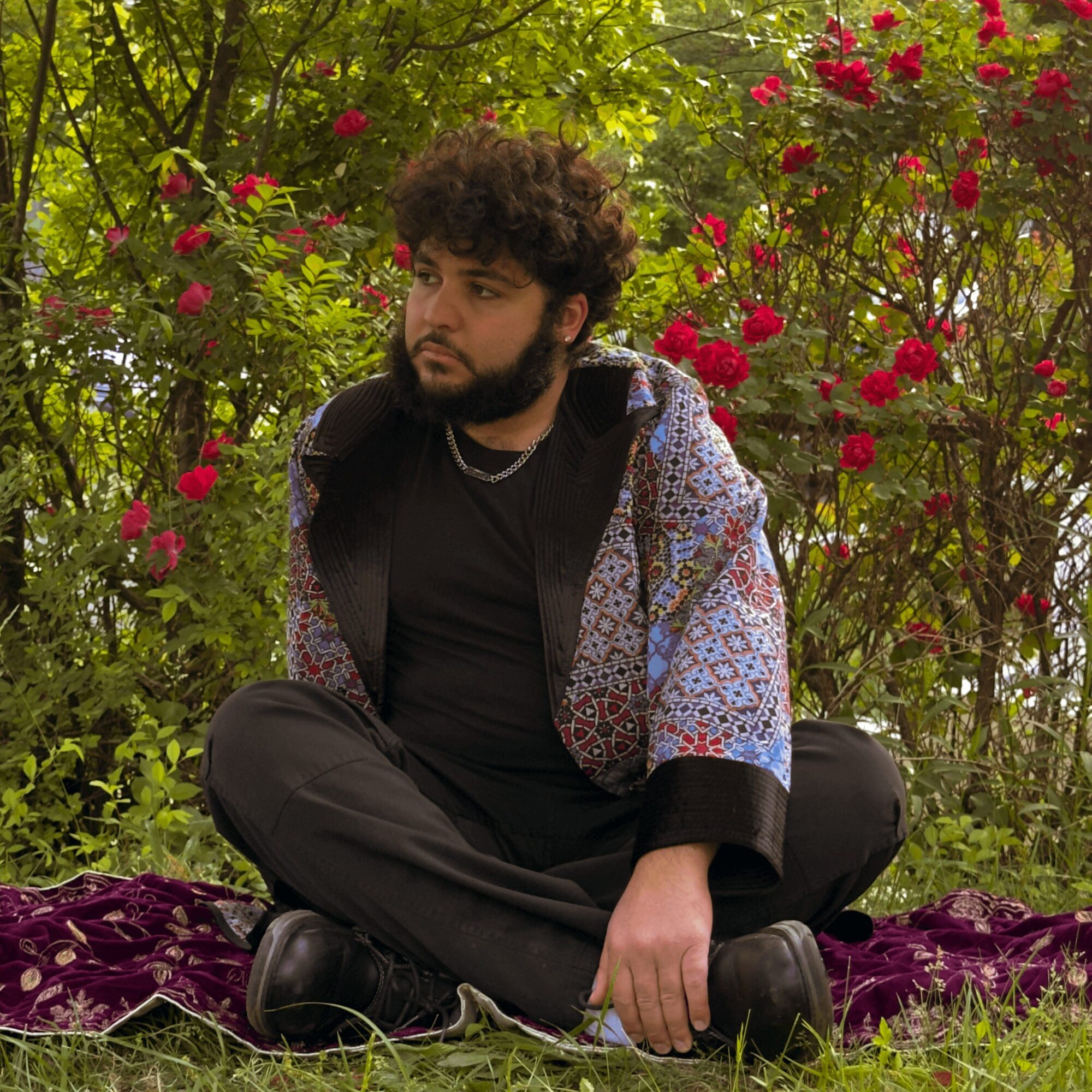 Young man with curly hair and beard sitting cross-legged outdoors among pink flowers and green foliage.