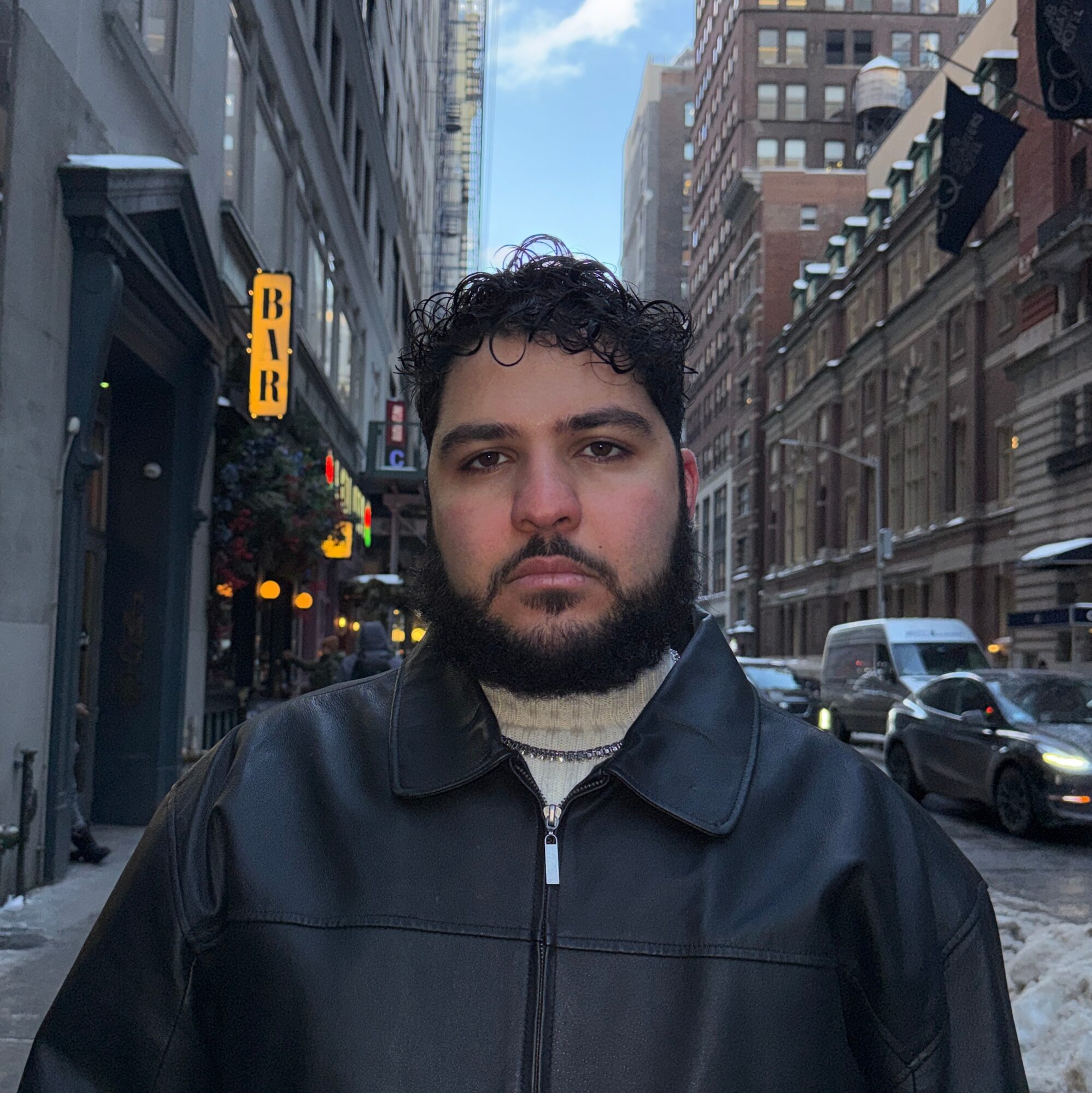 Young man with dark curly hair and beard wearing a black leather jacket on city street.