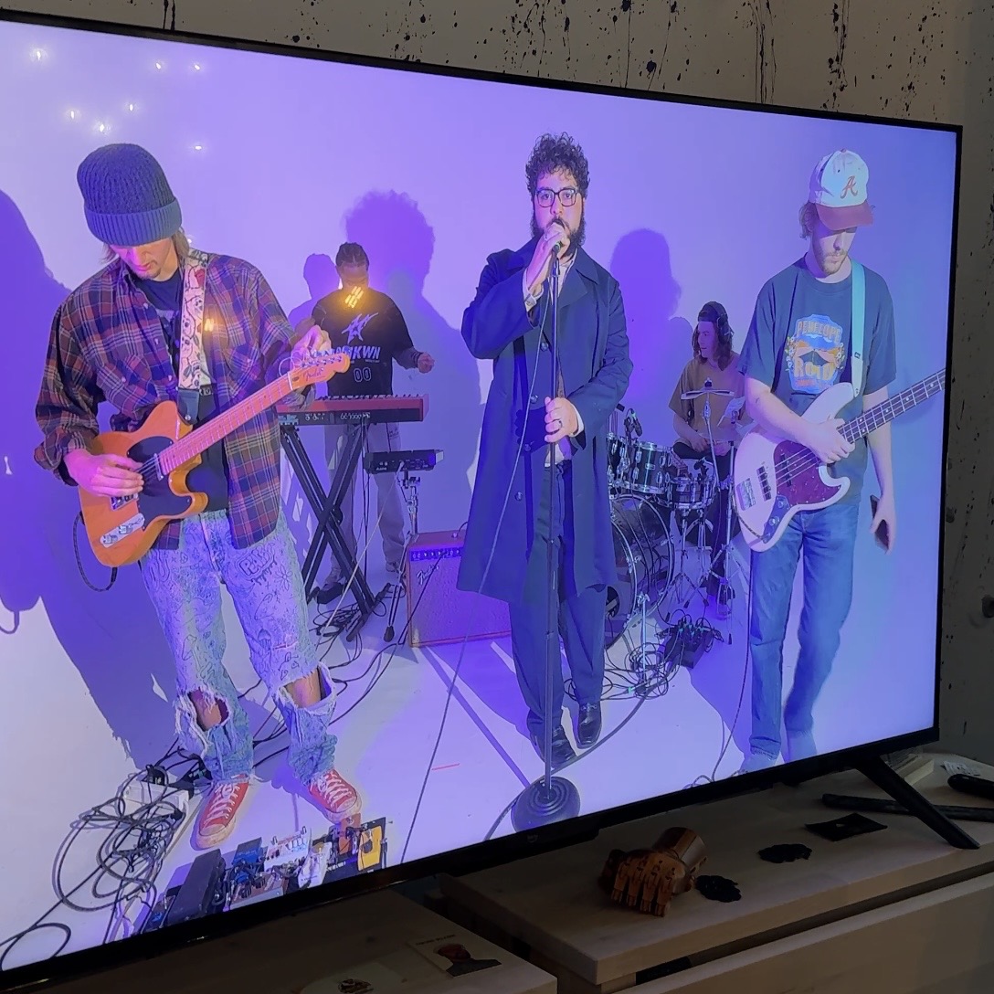 Band performing with four members playing guitar, keyboard, drums, and singing, against a purple background.