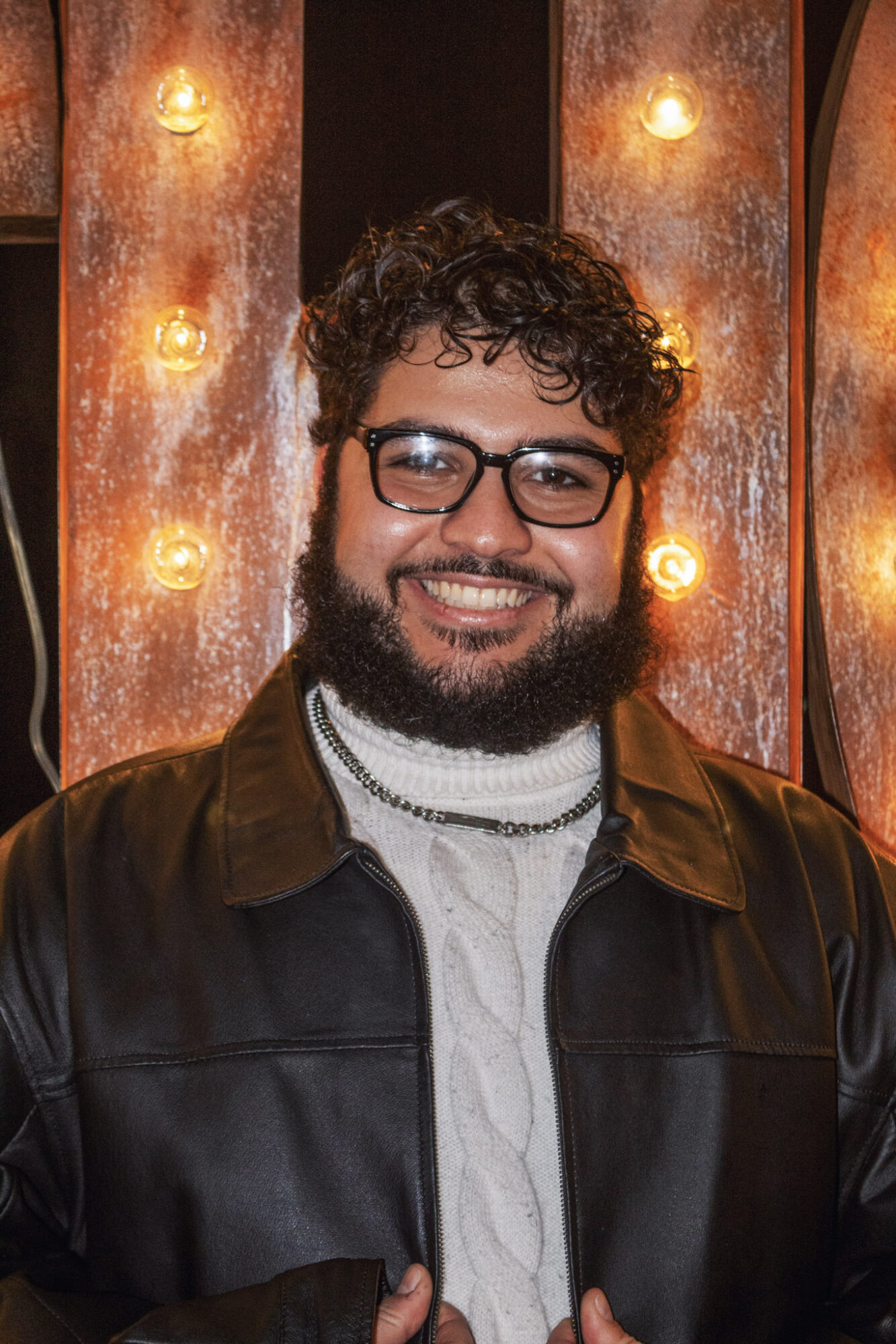 Man with curly hair, glasses, and beard smiling, wearing a leather jacket and white shirt, in a warmly lit setting.