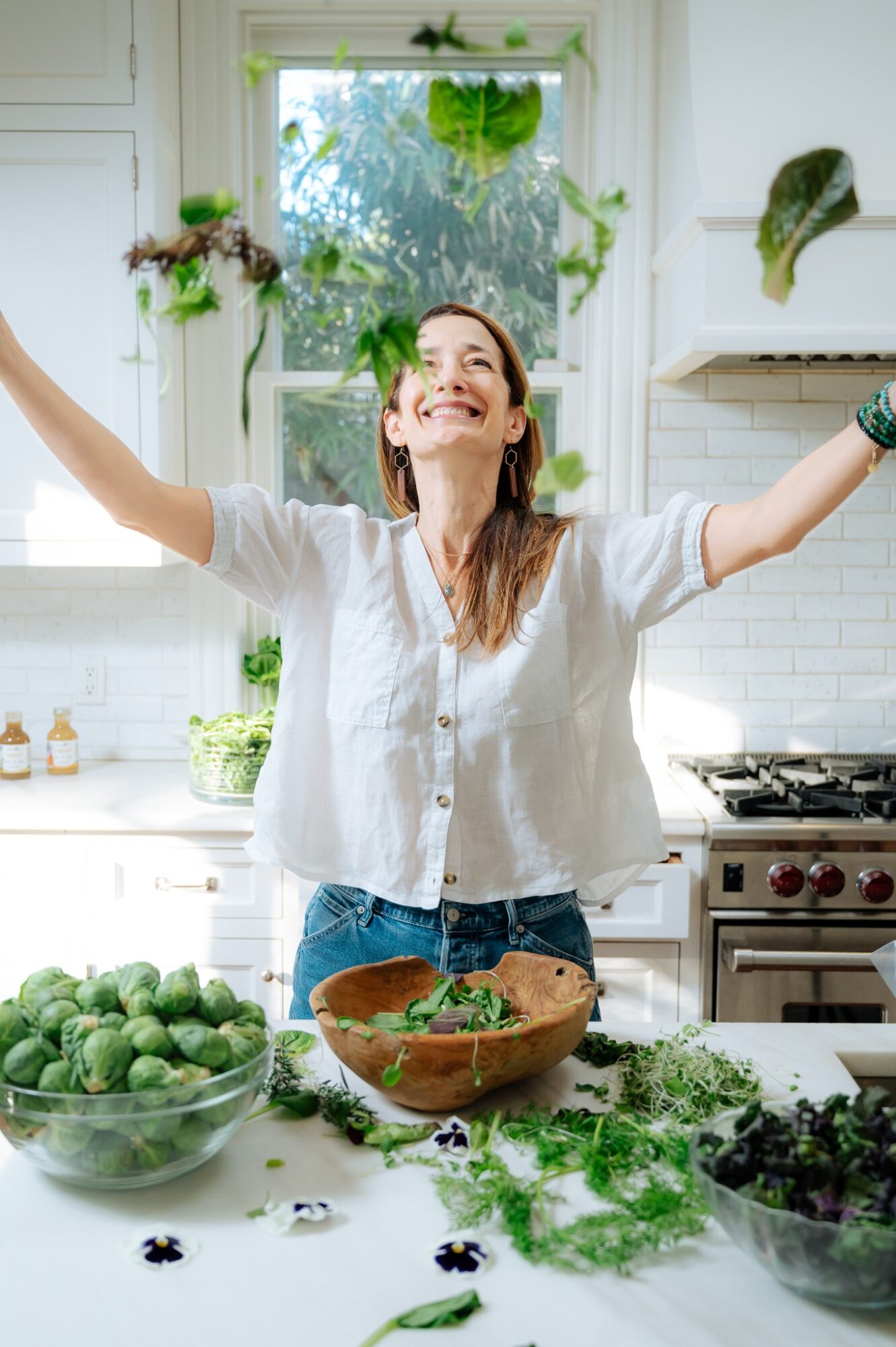 Woman in a kitchen with arms raised, smiling, surrounded by fresh vegetables and herbs, bright natural light from window.