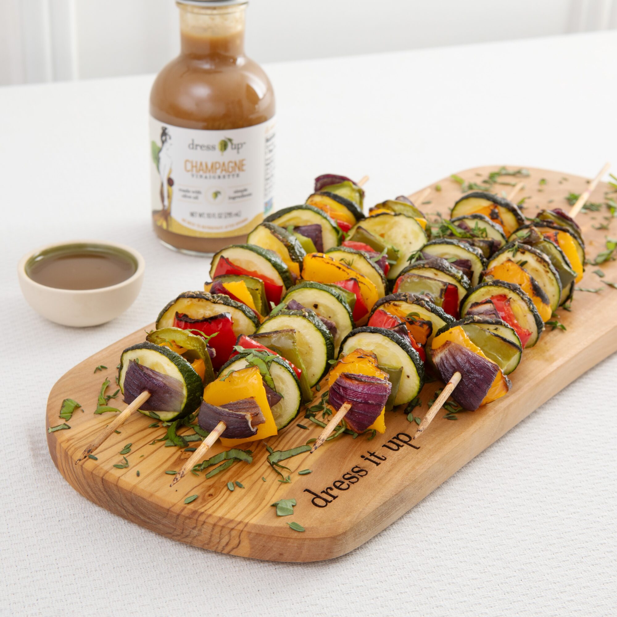 Skewers of colorful grilled vegetables on a wooden board with sauce and a bottle of drink in the background.