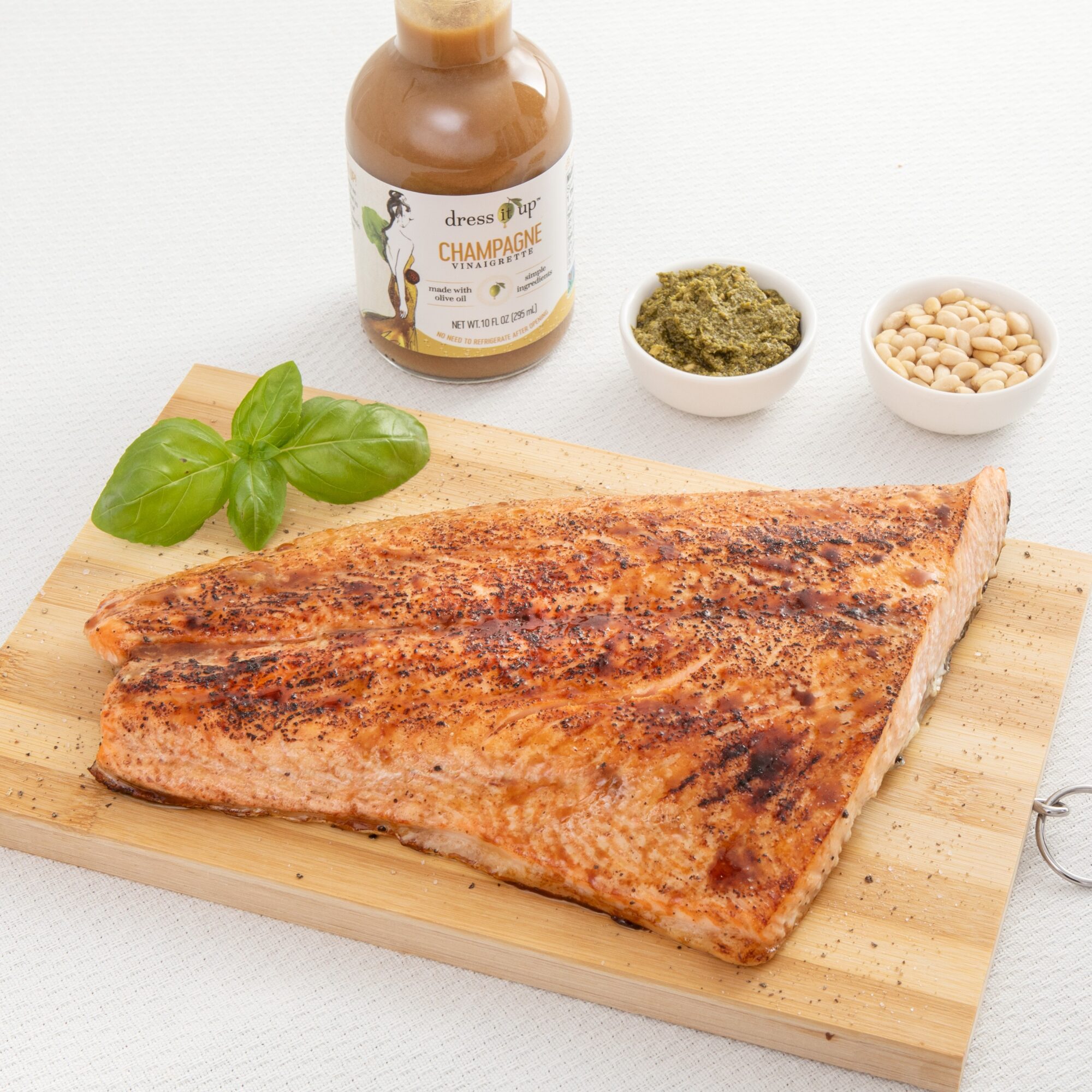 Grilled fish fillet on a wooden cutting board with basil leaves, a jar of sauce, and small bowls of pesto and beans, on a white surface.