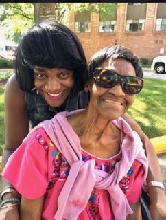 Two women smiling outdoors, one wearing sunglasses and a pink embroidered top, the other leaning over her shoulder.