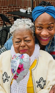 Two women, one elderly with gray hair and the other younger with a blue headwrap, smiling together outdoors.