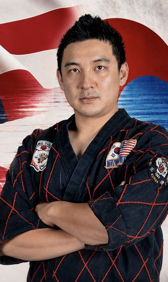 Man in a dark blue martial arts uniform with patches, arms crossed, standing in front of a red, white, and blue background.
