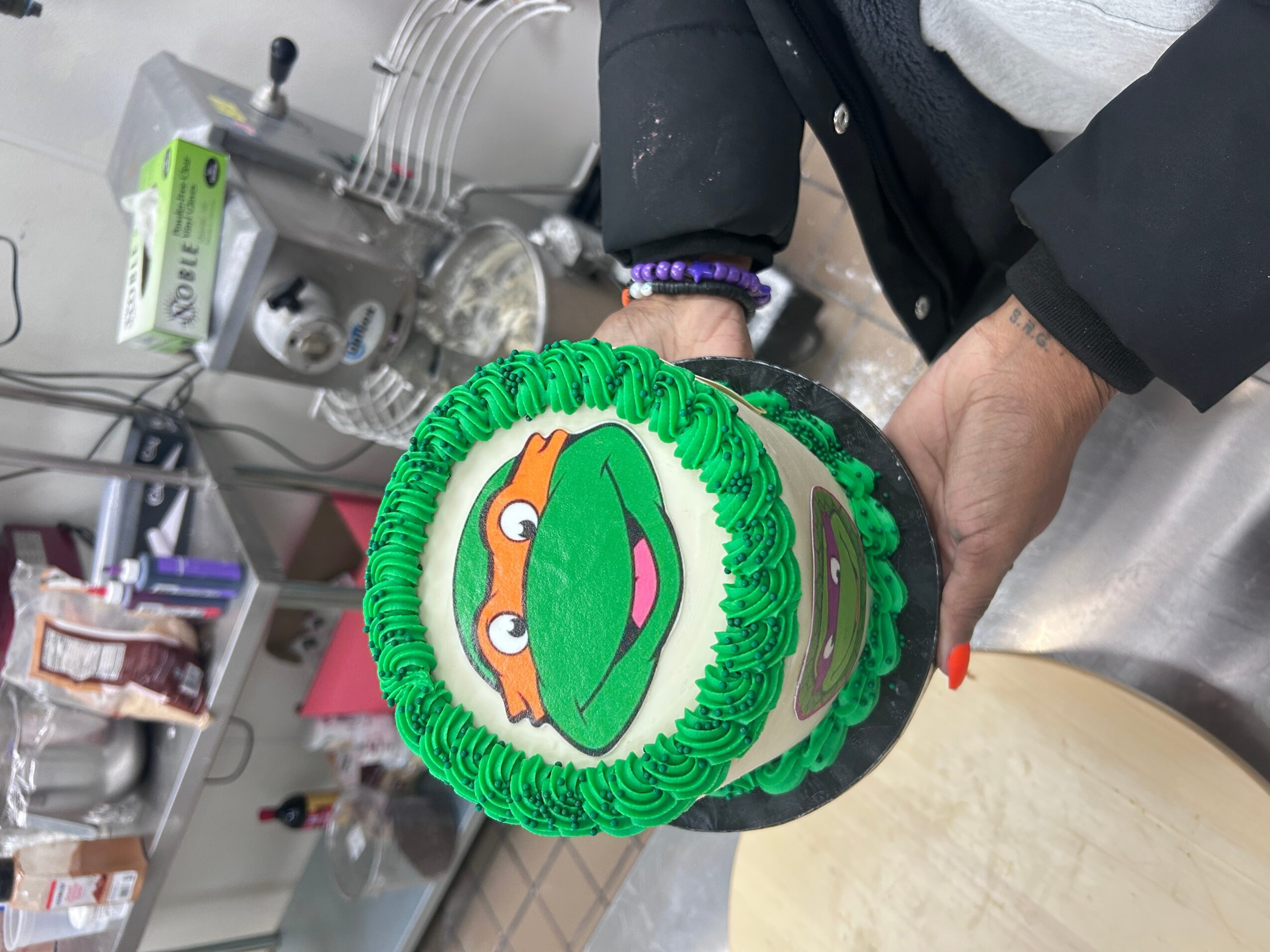 Person holding a cake with a cartoon face of a frog with orange eyes and a pink tongue, decorated with green frosting.