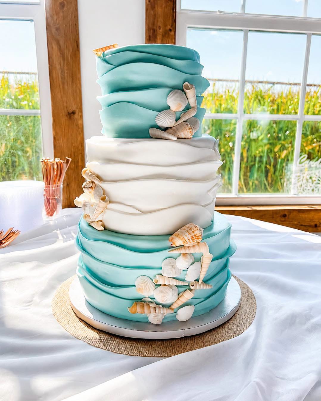 A three-tiered cake decorated with seashells and sea-themed elements, placed on a table near windows with a view of greenery.