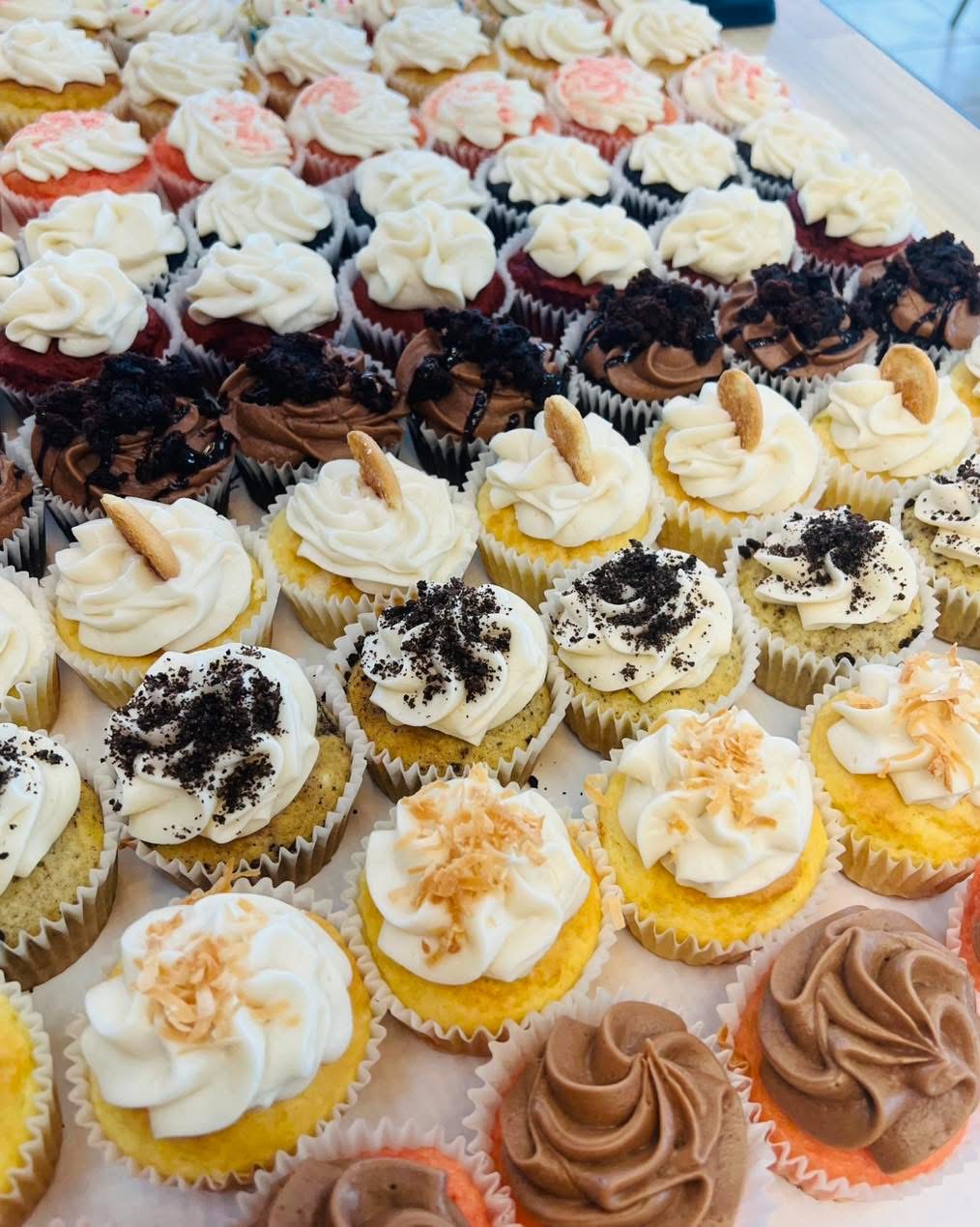 Assorted cupcakes with various toppings arranged on a table, featuring white, chocolate, and yellow frosting, some with sprinkles or crumbs.