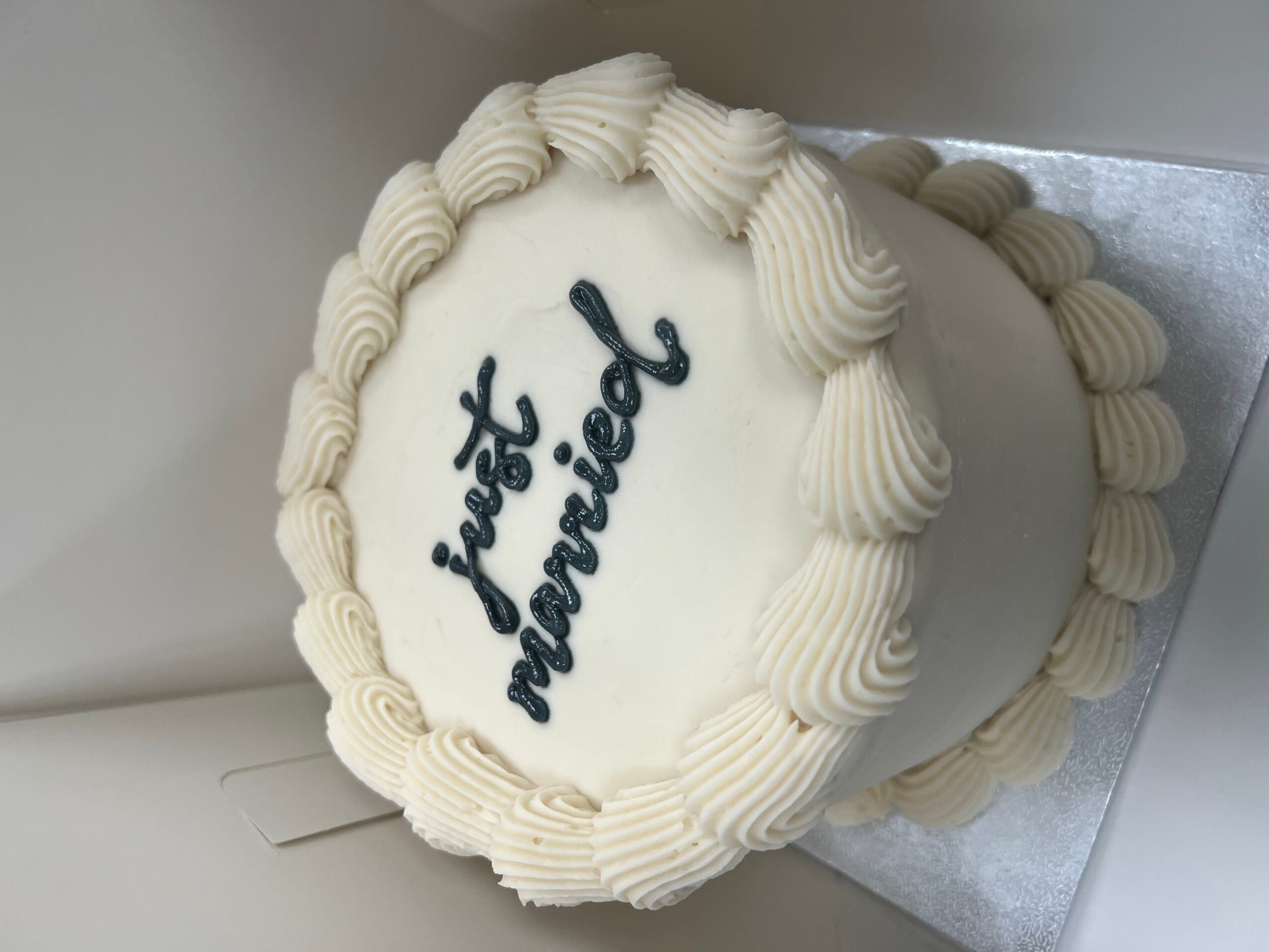 Round wedding cake with white frosting and piped border, decorated with the words 'just married' in dark icing.