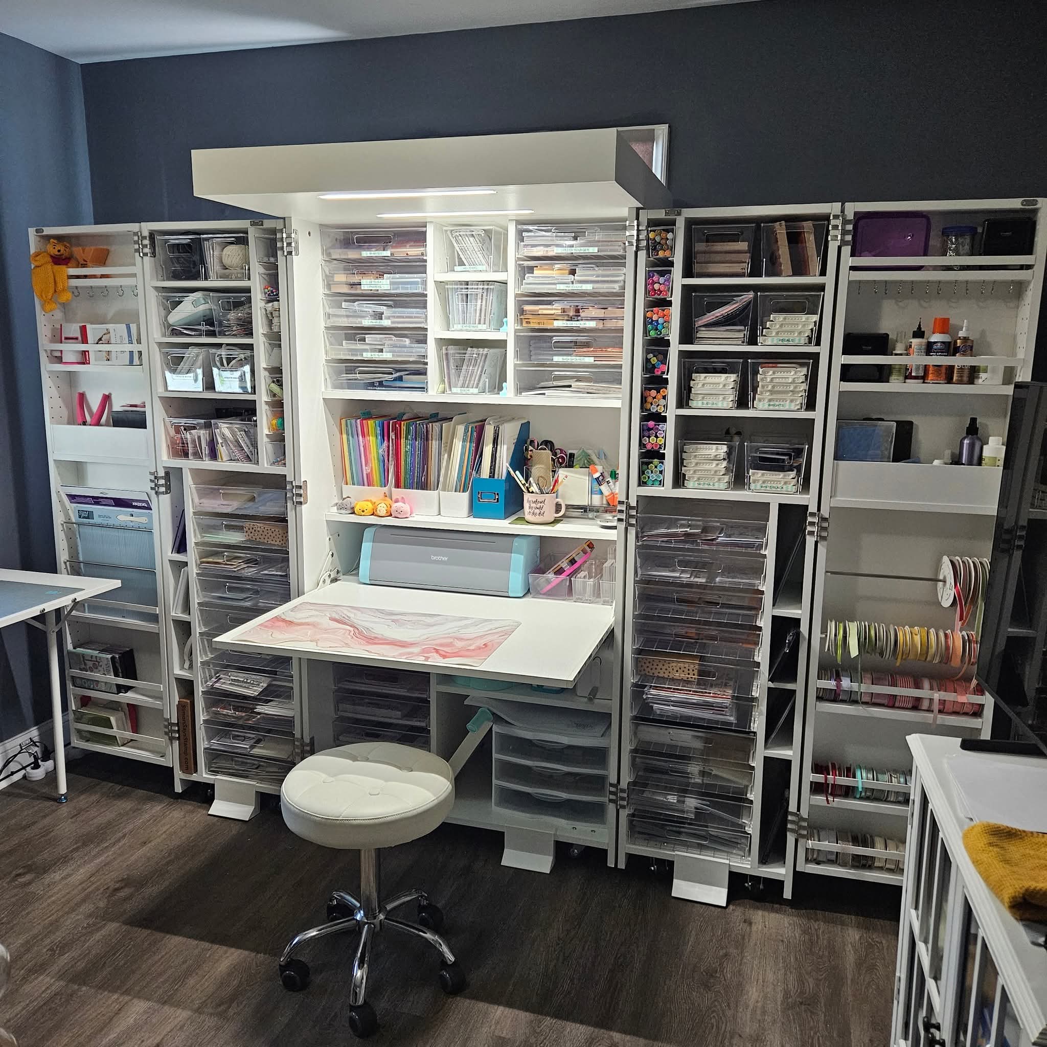 Organized craft supply storage with shelves filled with books, containers, and craft materials, in a room with dark flooring and blue walls.