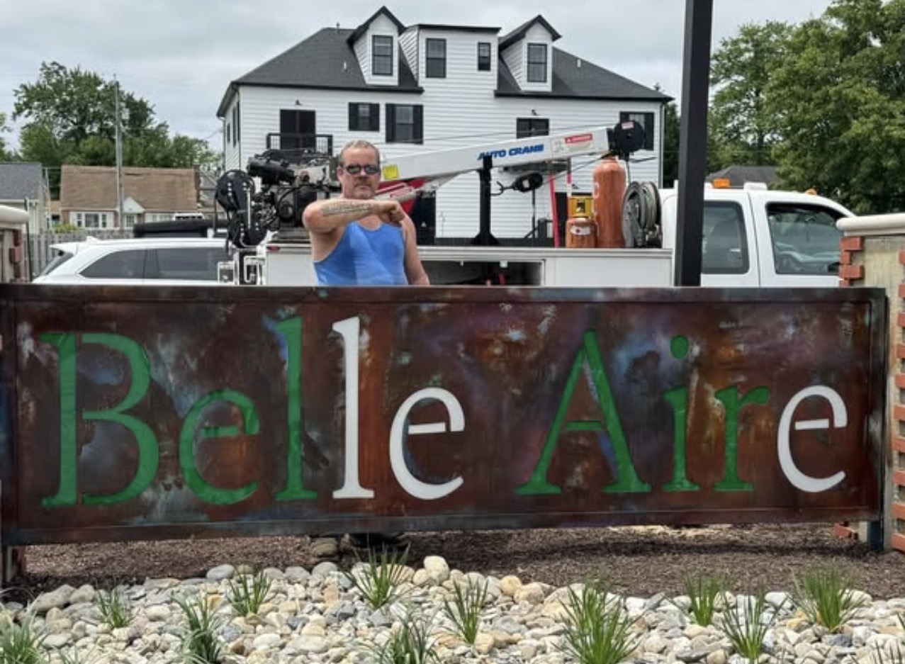 Person operating camera behind a sign that reads 'Belle Aire' with a house and trees in the background.