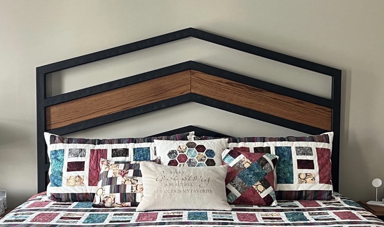 Bed with a geometric headboard, pillows, and a quilted bedspread, against a plain wall.