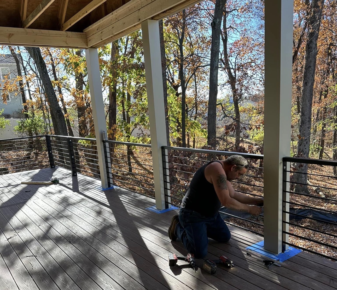 Person kneeling on a deck installing a railing, with trees and sunlight in the background.