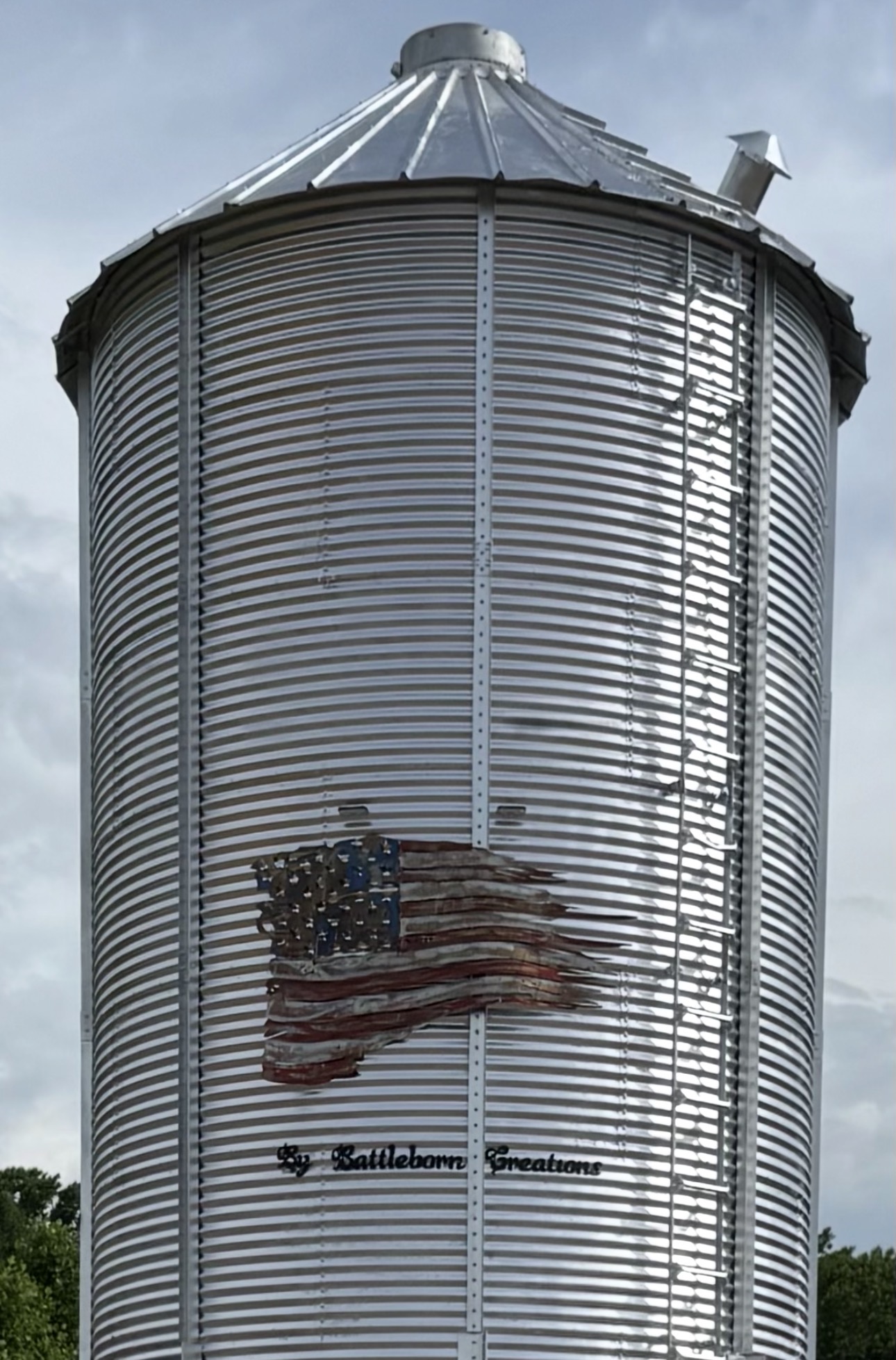 Silver cylindrical grain silo with an American flag and text on it, against a cloudy sky.