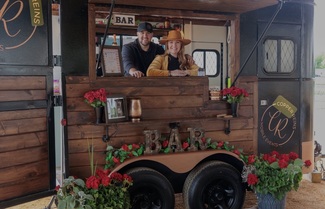 Two people in a bar setup with flowers and decorations, smiling behind a wooden counter, outdoors.