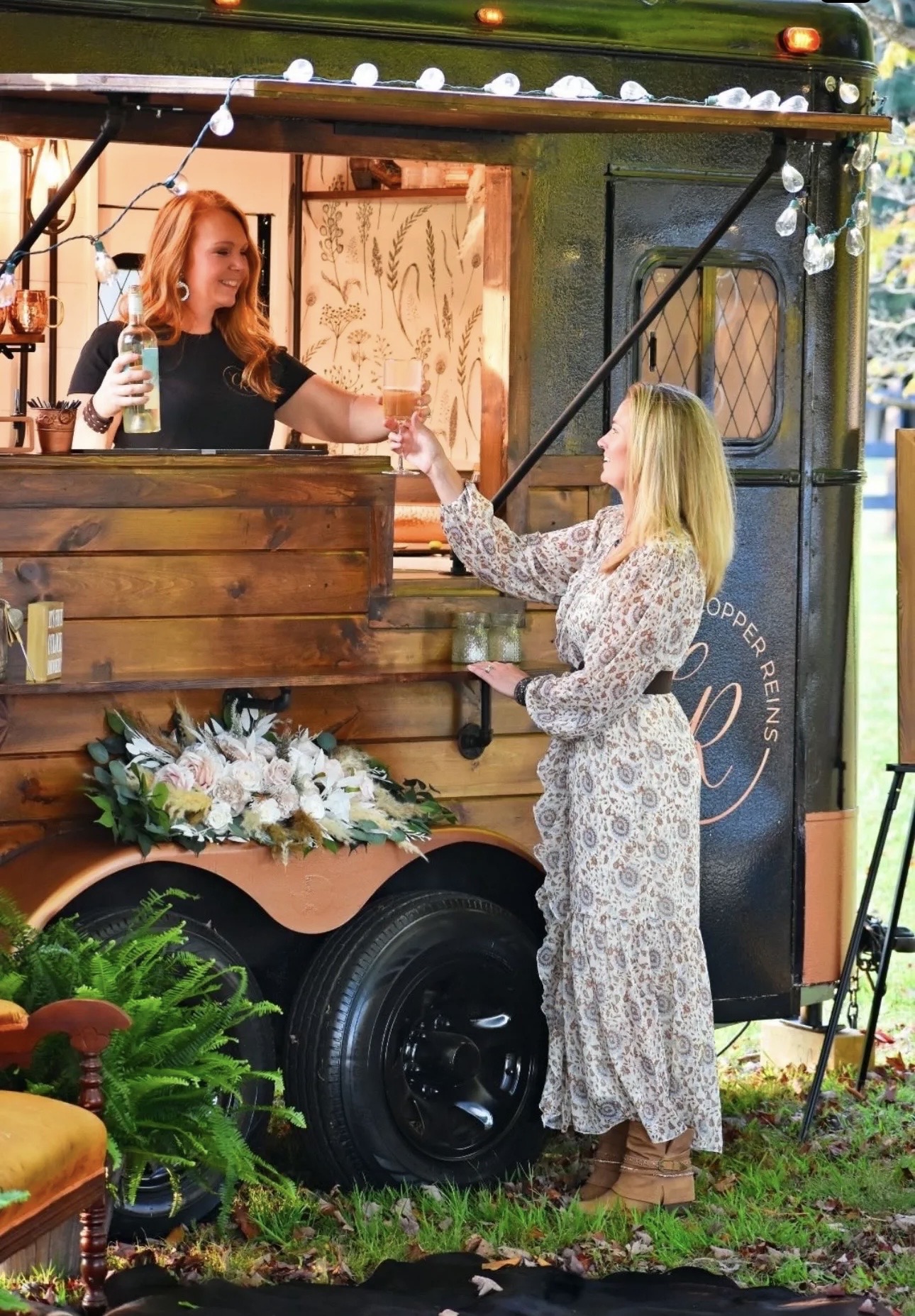 Two women exchanging a drink and a gift at a decorated outdoor event, with a floral arrangement nearby.