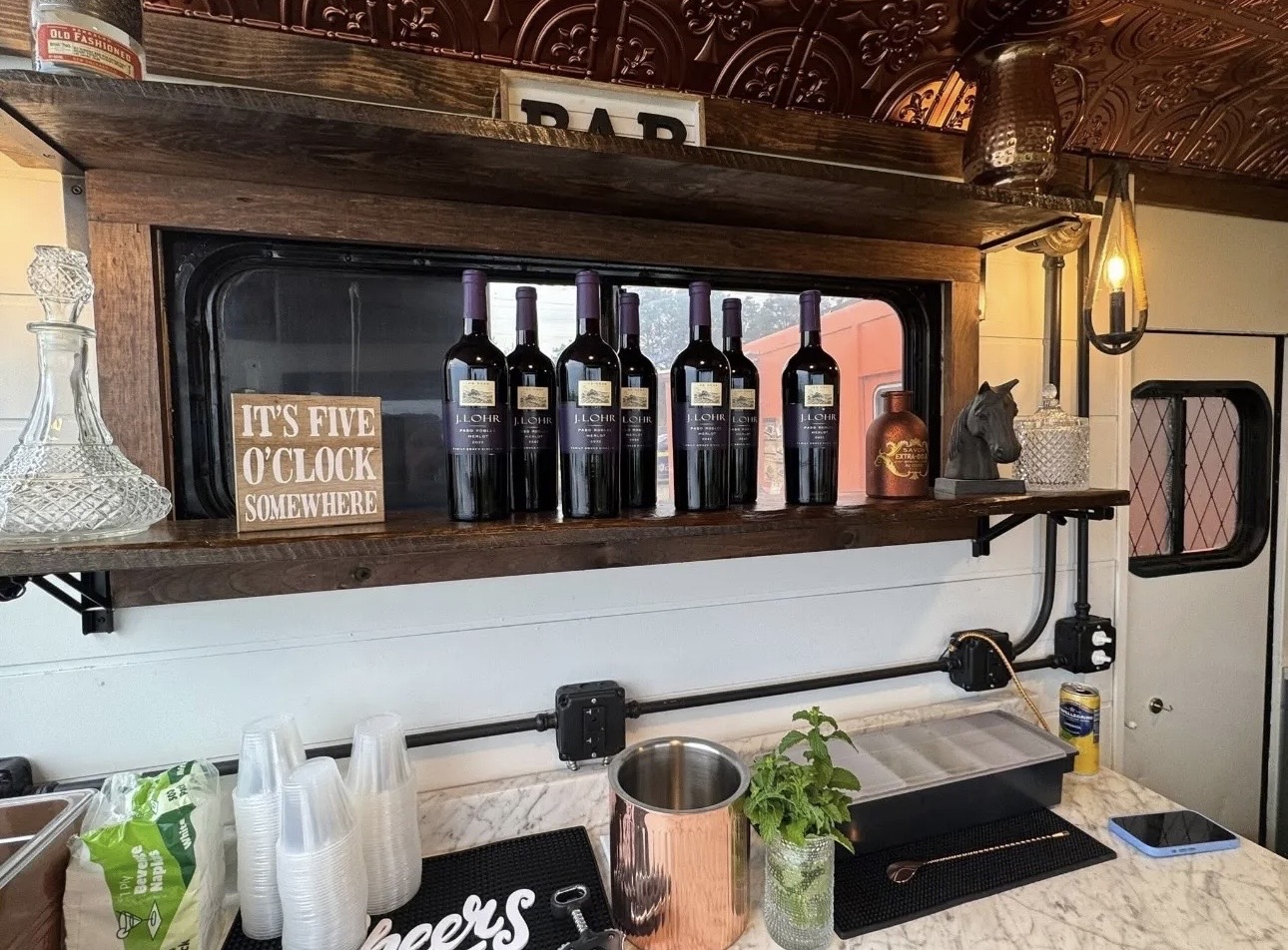 Shelf with wine bottles, a sign, and decorative items above a counter with cups and plants.