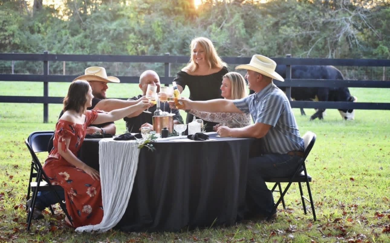 Group of five people having a meal outdoors at a round table with a black tablecloth, in a grassy area with trees and a fence.