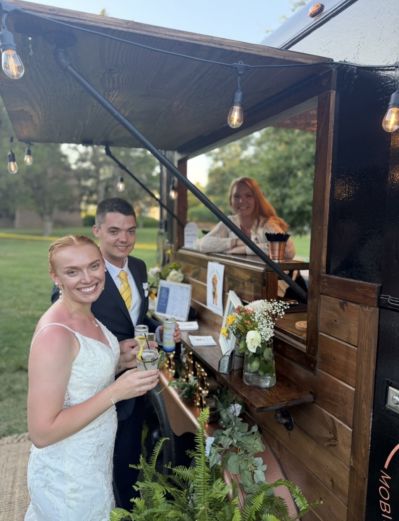 A smiling woman in a white dress and a man in a suit stand at a food truck window outdoors, holding drinks, with a woman inside the truck. Flowers and greenery decorate the truck window.