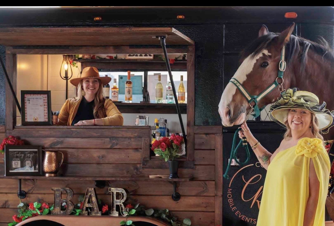 Woman in yellow dress and hat with a horse at a wooden bar setup, decorated with flowers and signs.