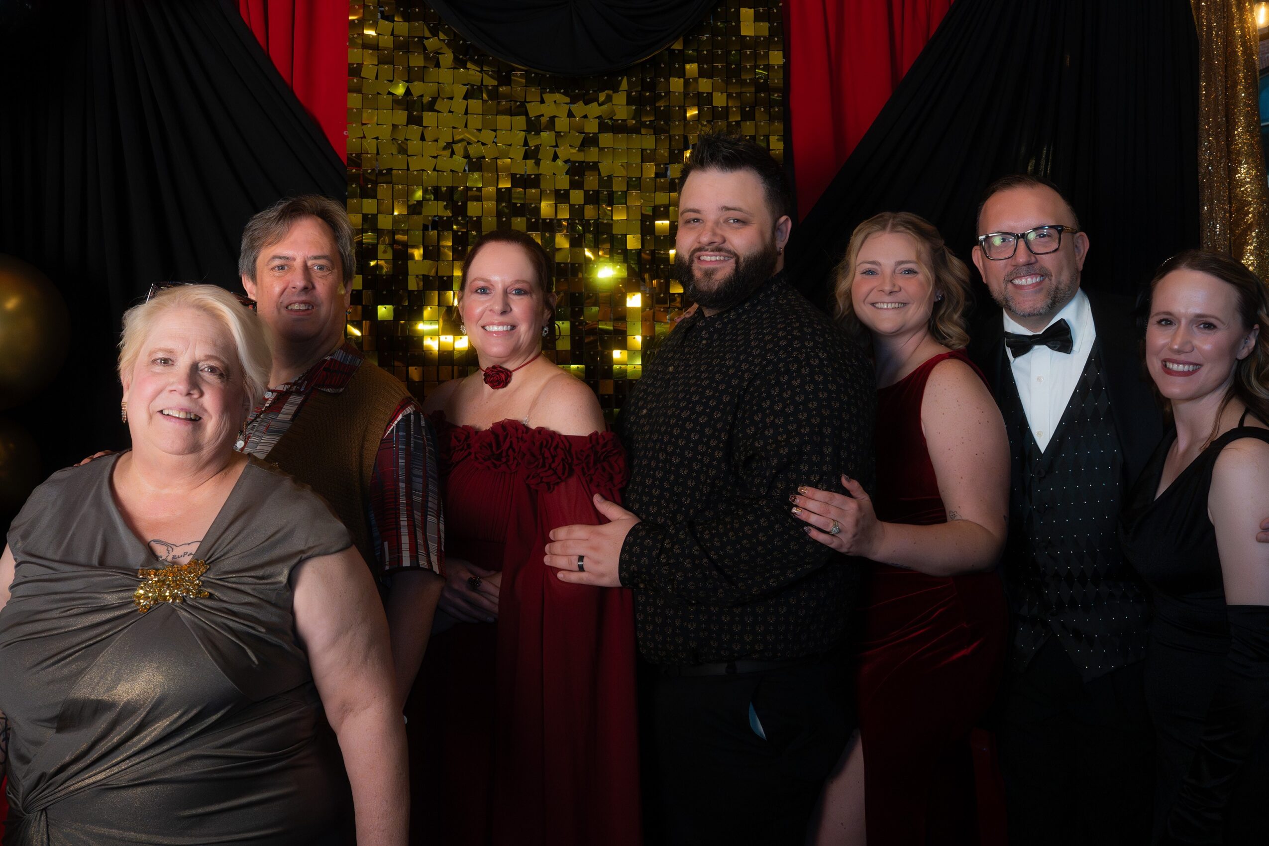 Group of seven people dressed in formal attire, smiling in front of a decorated backdrop with red and black curtains and gold sequins.