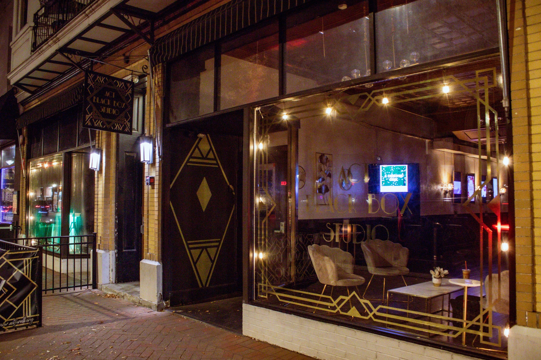 View of a storefront with large glass windows and a black door, decorated with gold geometric patterns, illuminated interior, and seating visible inside.
