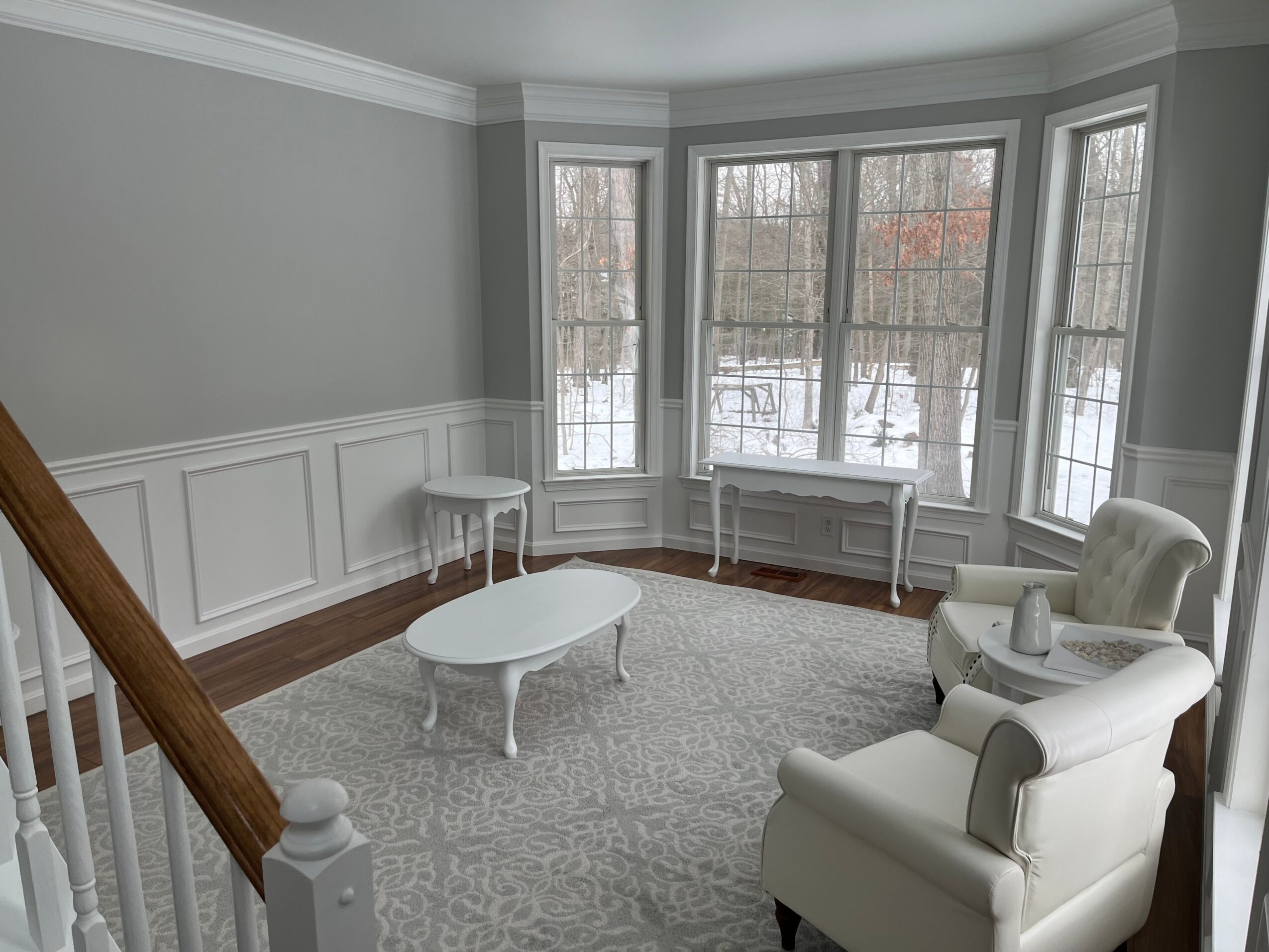 Living room with large bay window, white armchair, coffee table, and gray walls with white wainscoting.
