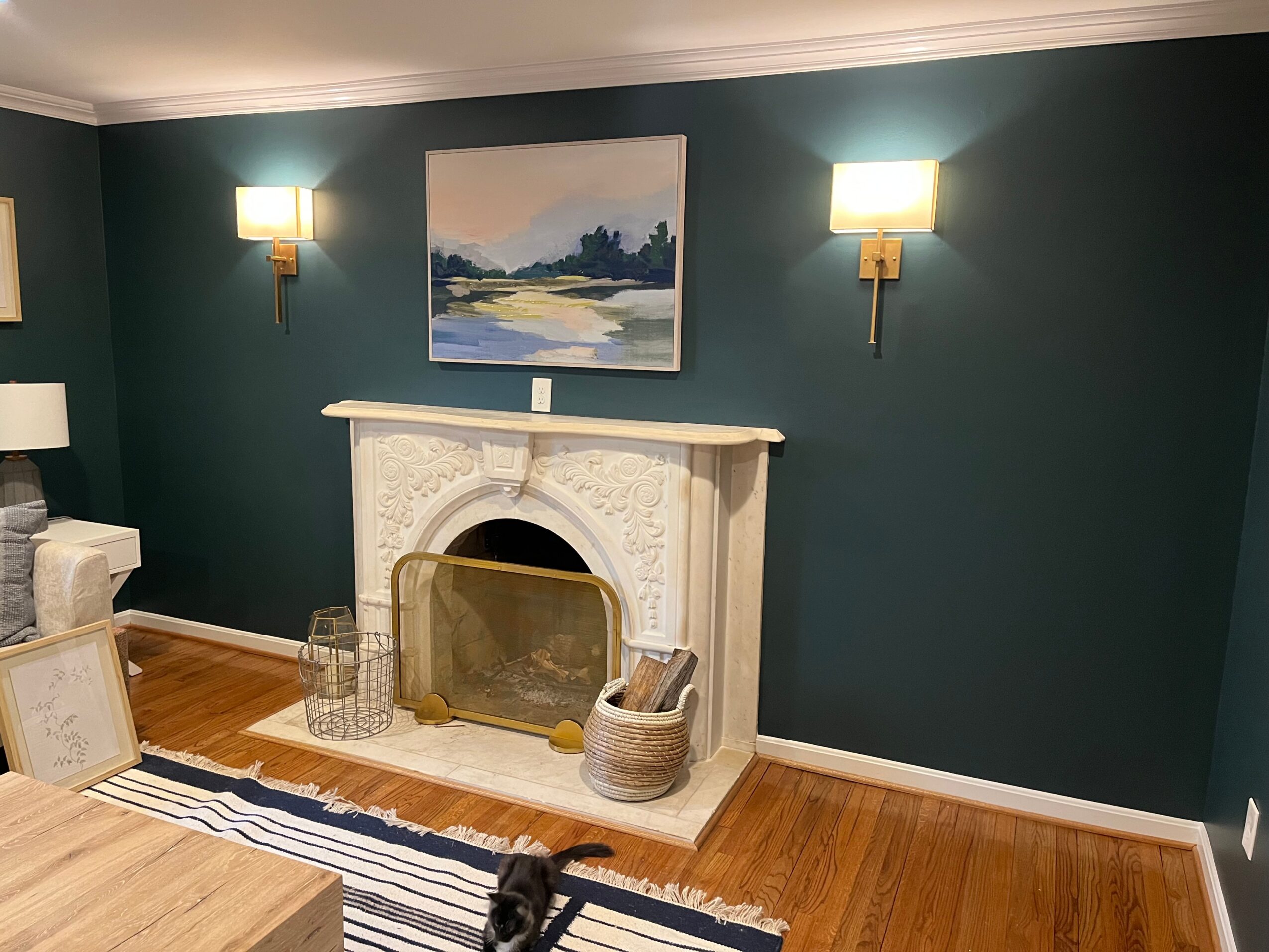 Living room with teal wall, white fireplace, two wall sconces, painting above fireplace, and hardwood floor.