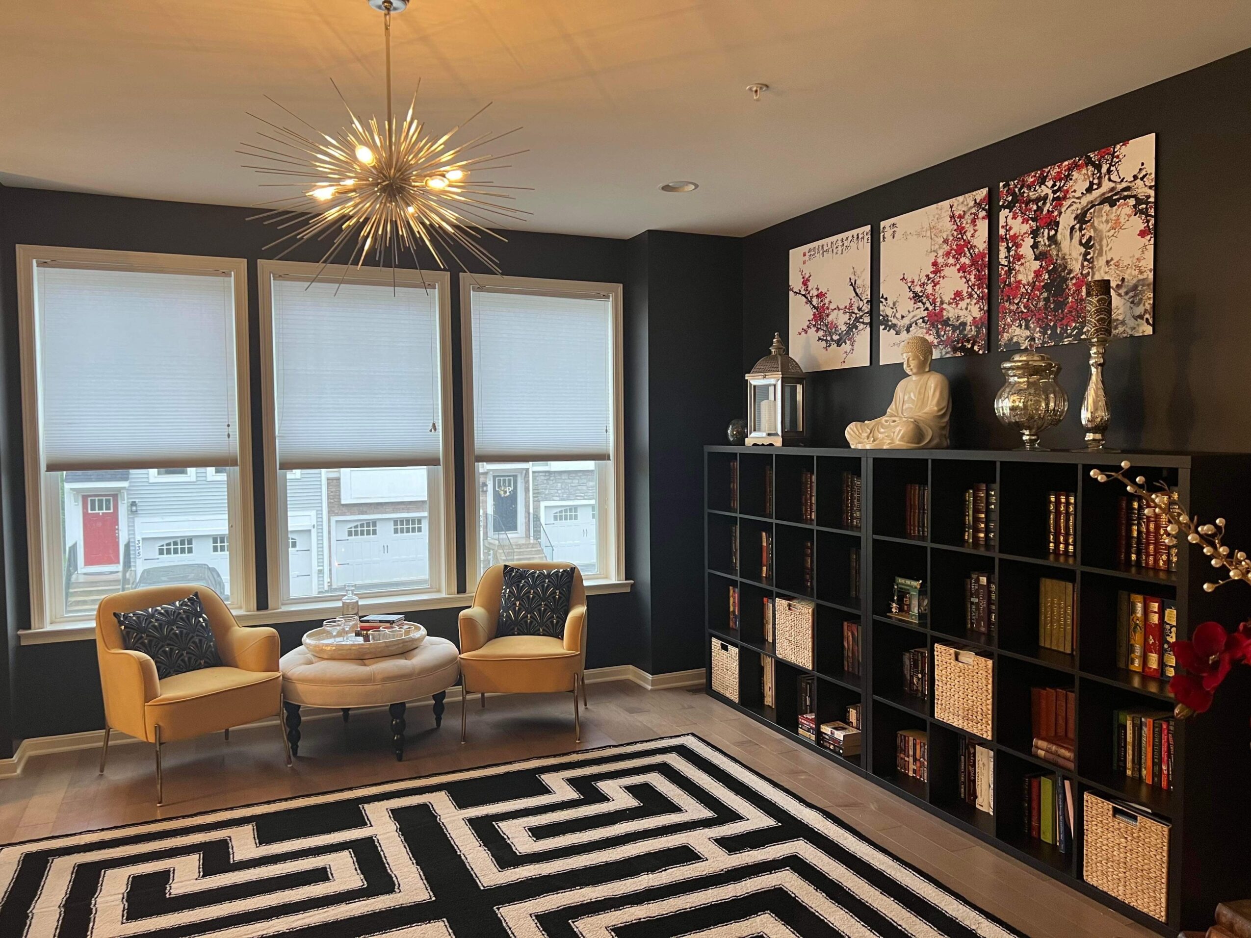 Living room with three large windows, two yellow armchairs, a patterned rug, and a black bookshelf with decorative items.