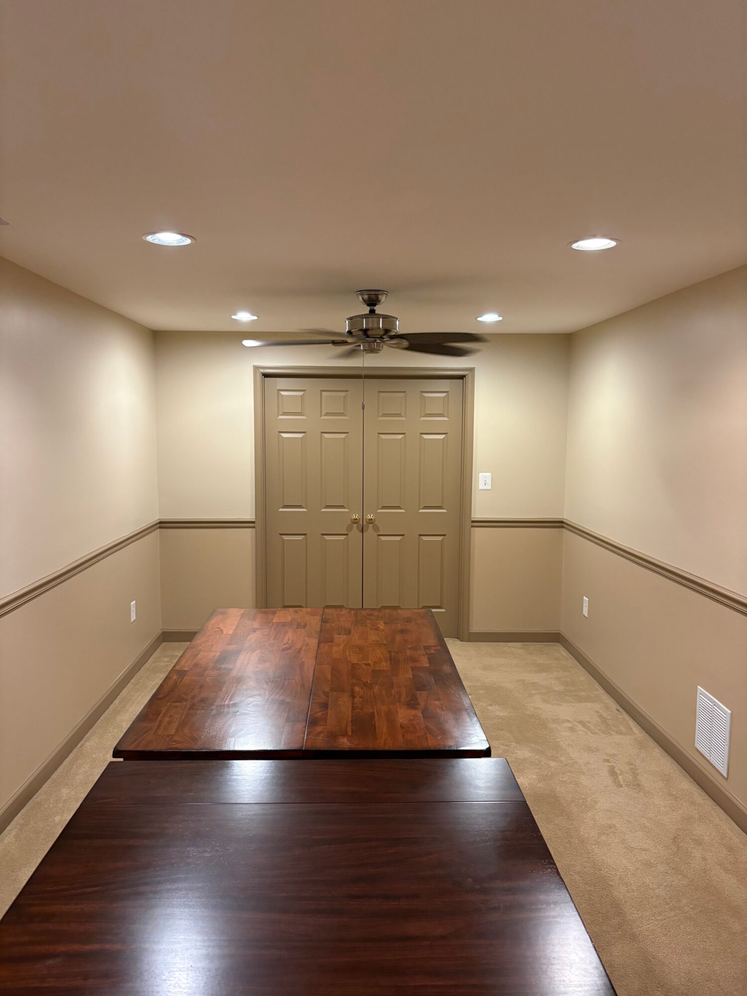 Upright view of a room with beige walls, a ceiling fan, and a closed double door, with a dark wooden floor and carpeted area.