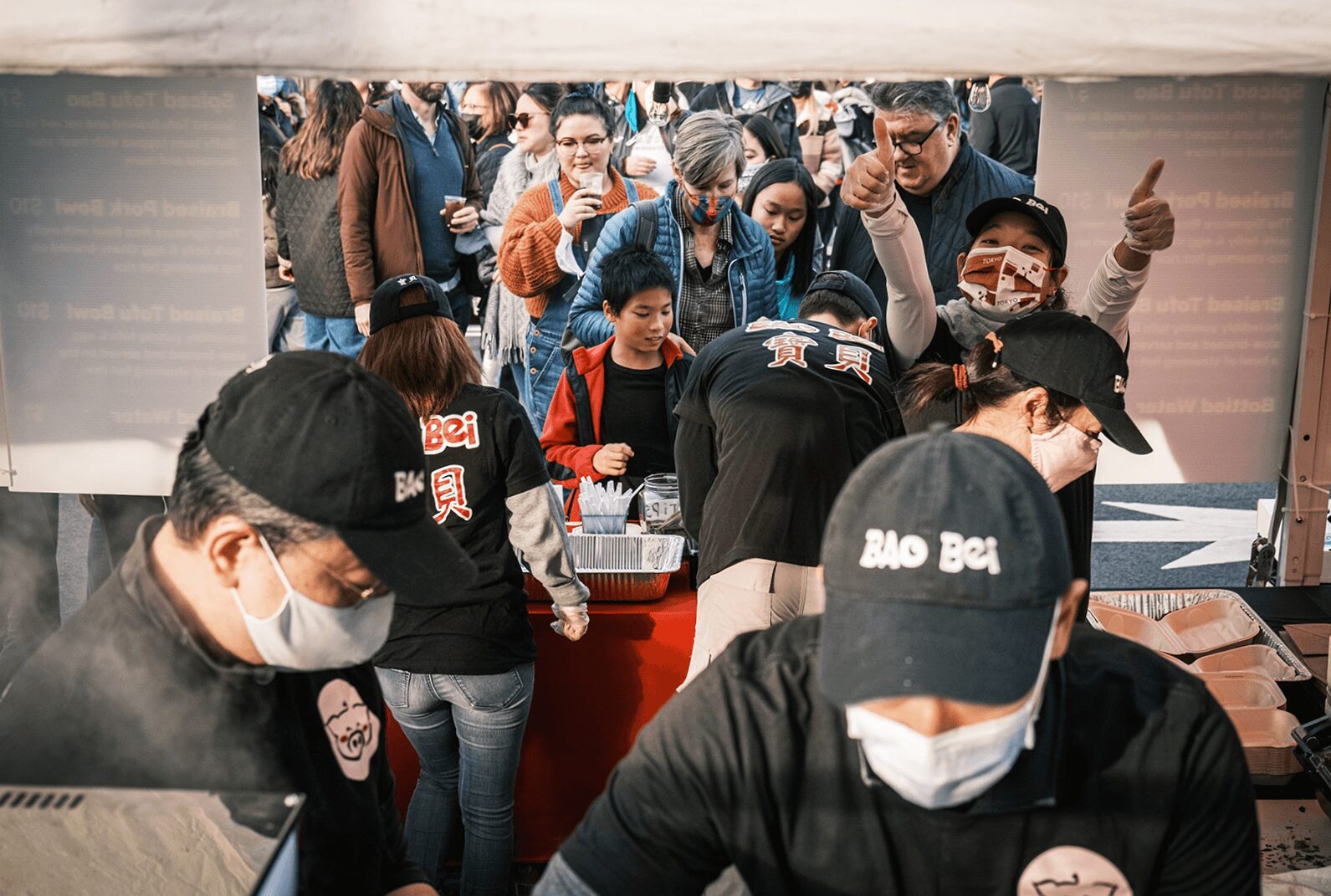 People gather around a booth, some wearing masks and hats, with a crowd behind them.
