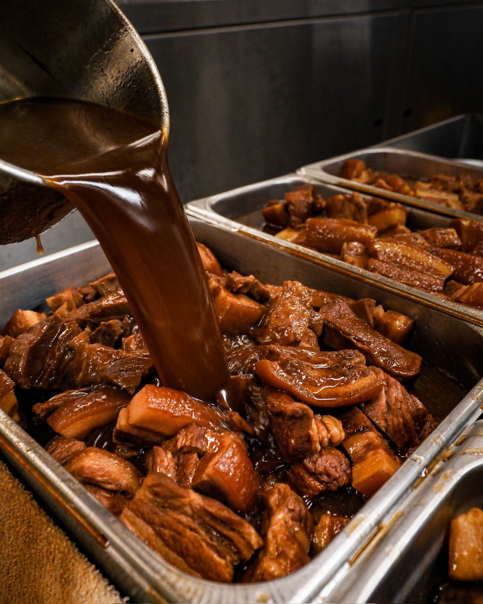 Pouring dark gravy over cooked meat and vegetables in a metal tray.