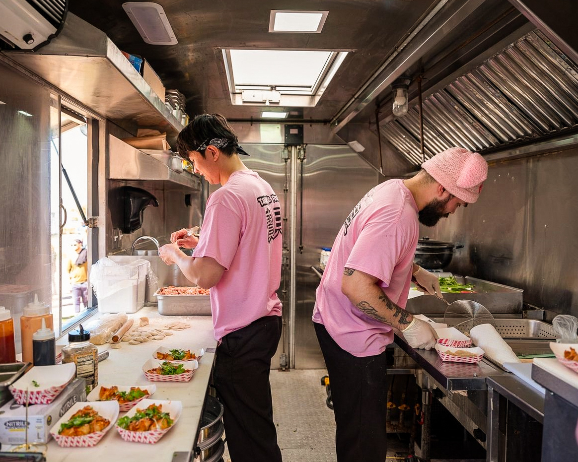 Two people in pink shirts preparing food in a commercial kitchen with stainless steel surfaces and cooking equipment.