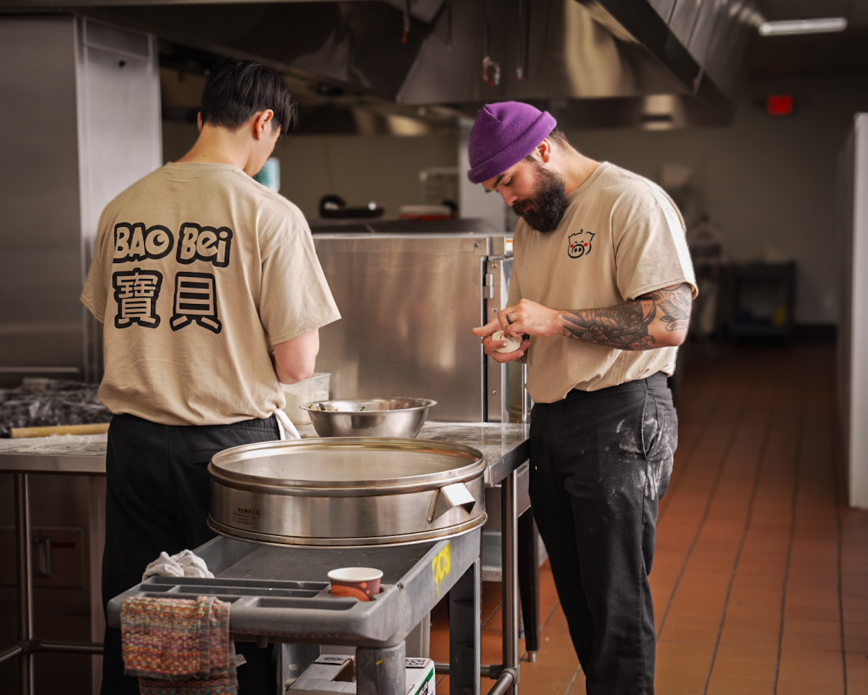 Two chefs in a kitchen, one with a purple beanie and tattoos, the other with Asian features, preparing food.