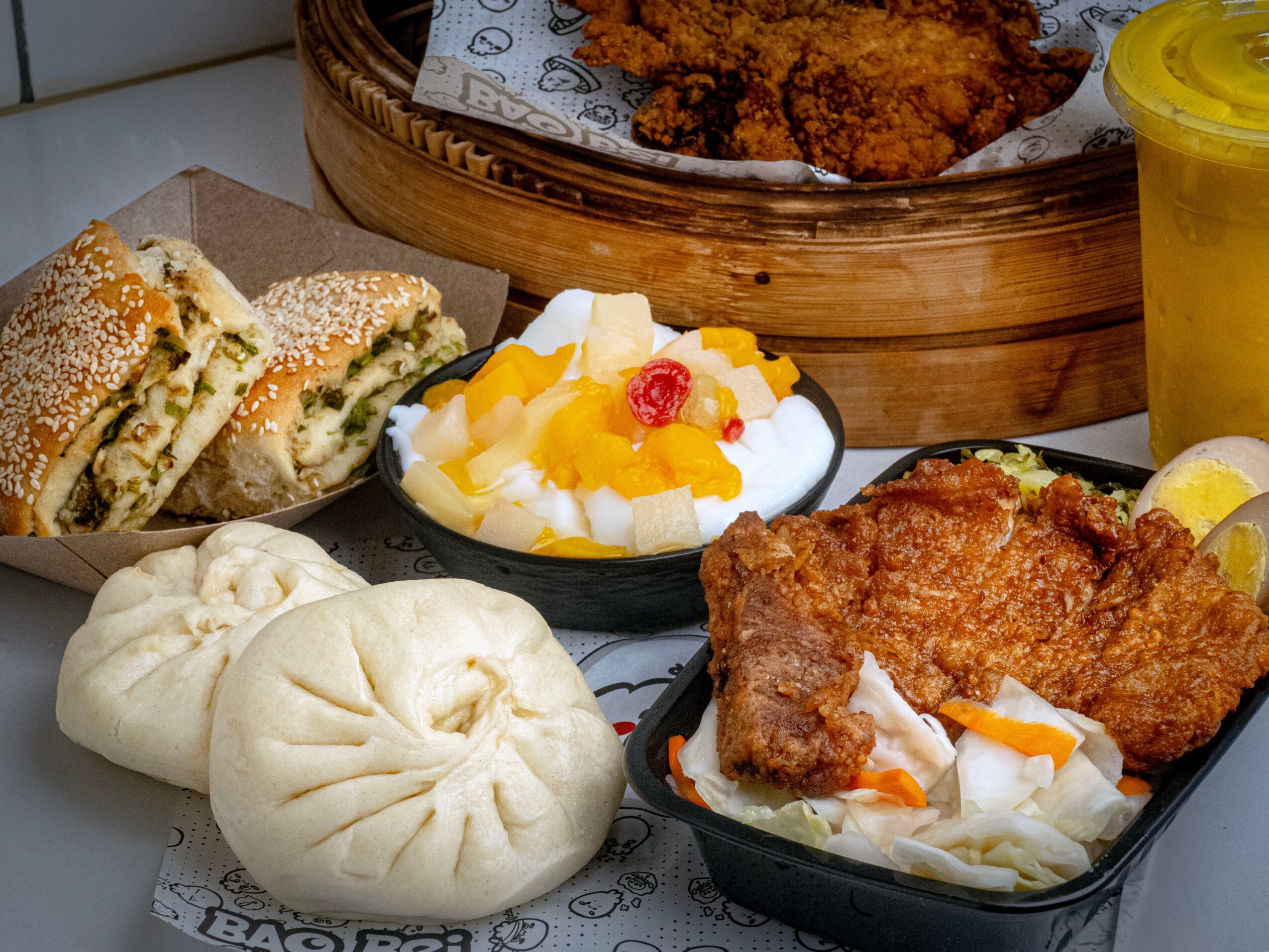 Assorted Asian dishes including steamed buns, fried chicken, rice with vegetables, and a drink, arranged on a table.