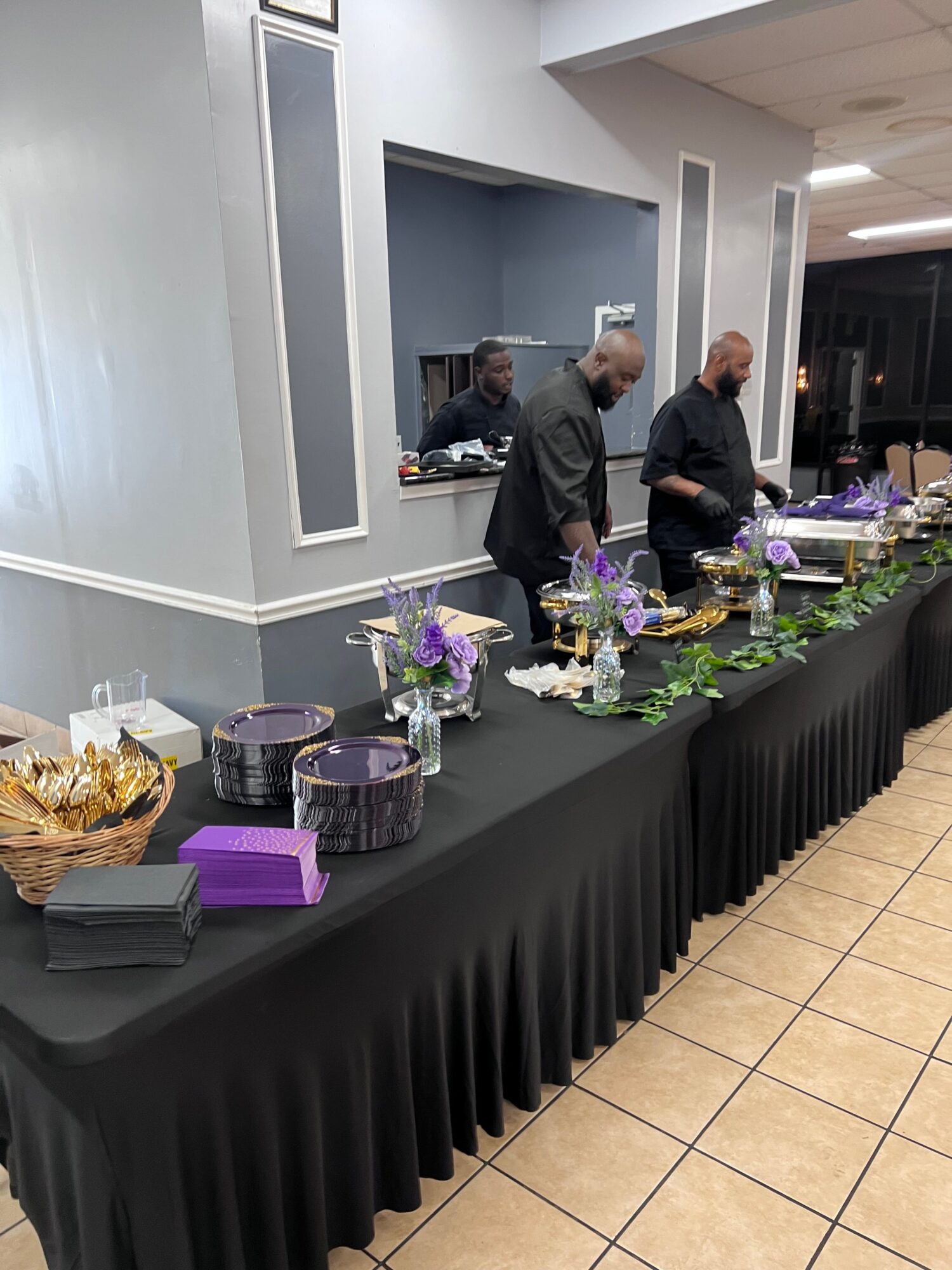 People standing at a table with purple and gold decorations, in a room with tiled floor and white walls.