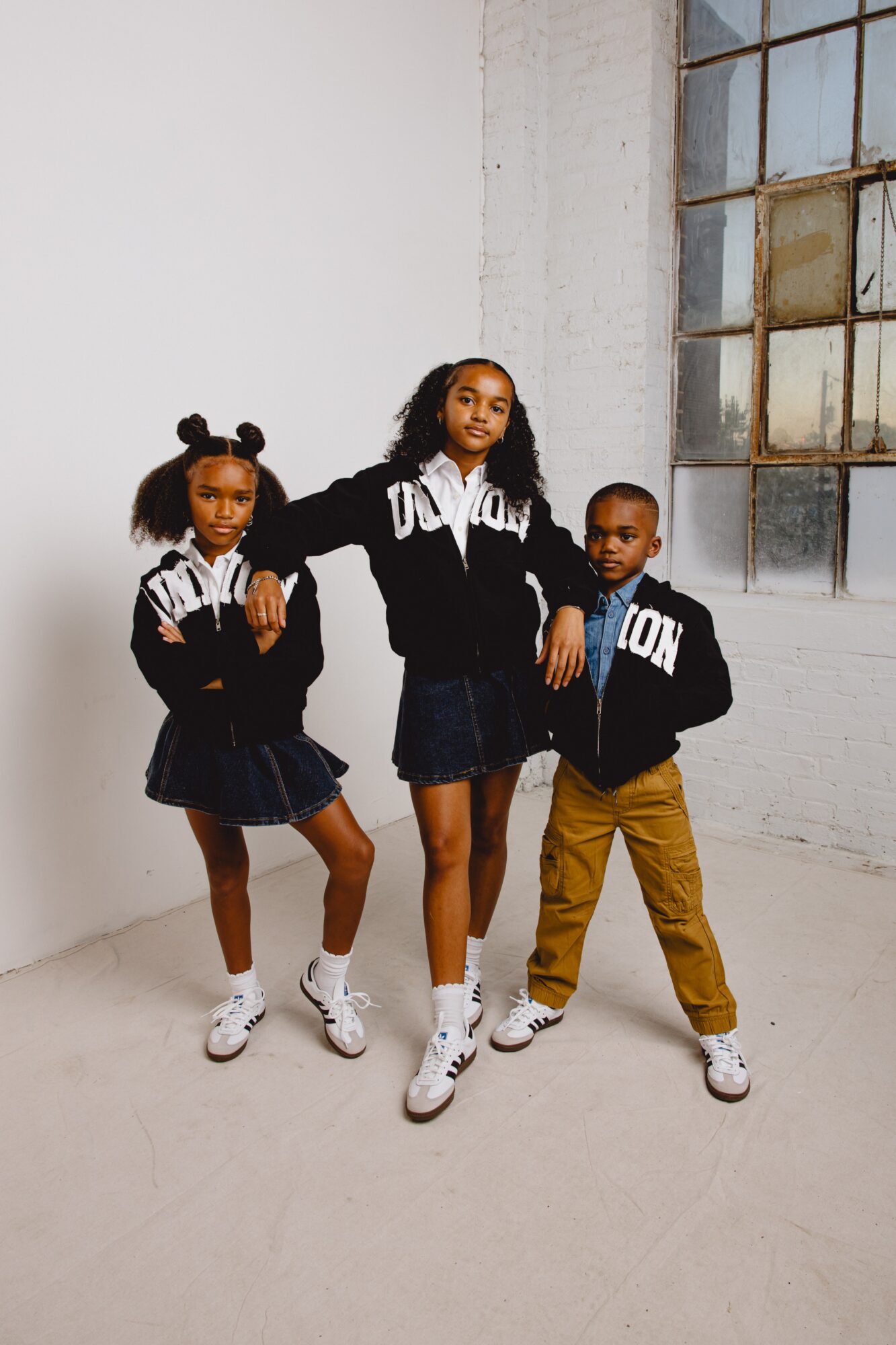 Three children in black and white hoodies and casual clothing pose indoors near a large window.