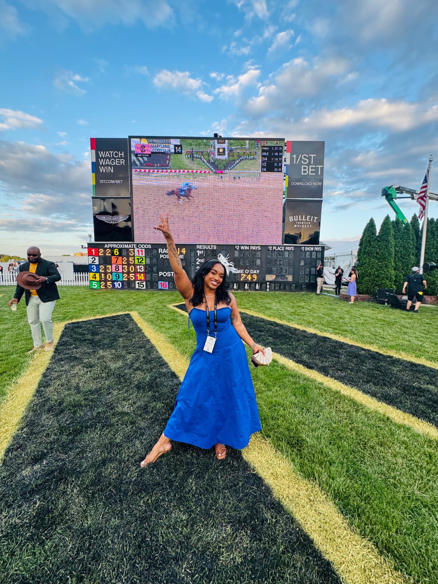 Woman in a blue dress celebrating outdoors in front of a large screen, with a partly cloudy sky above.