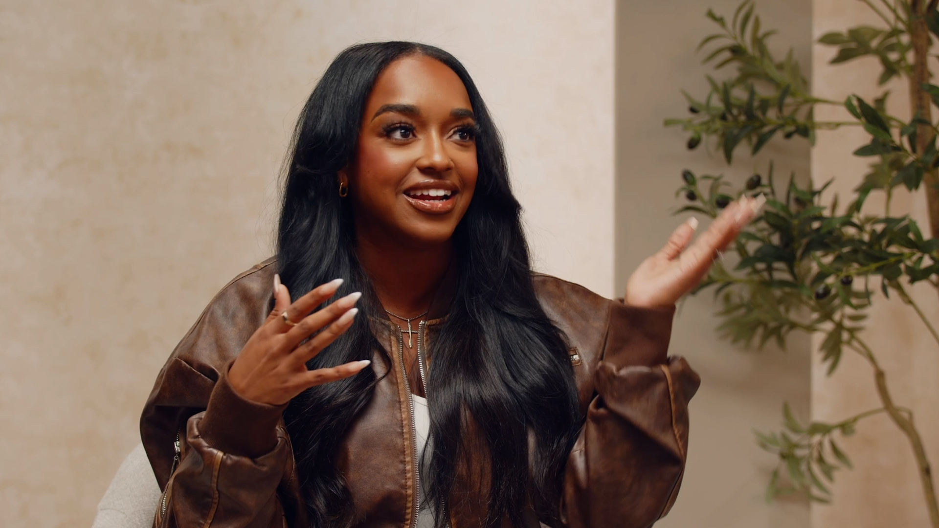 Woman with long black hair smiling and gesturing with her hands indoors, with a plant in the background.