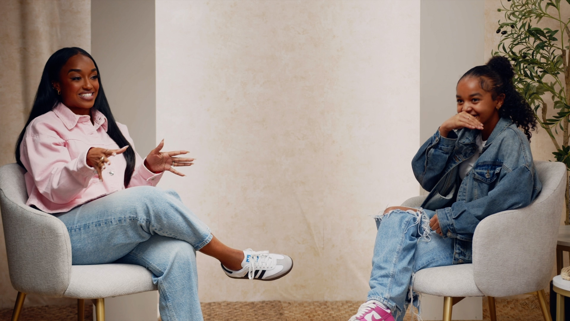 Two women sitting in chairs, smiling and talking, one wearing a pink shirt and jeans, the other in a denim jacket and jeans.