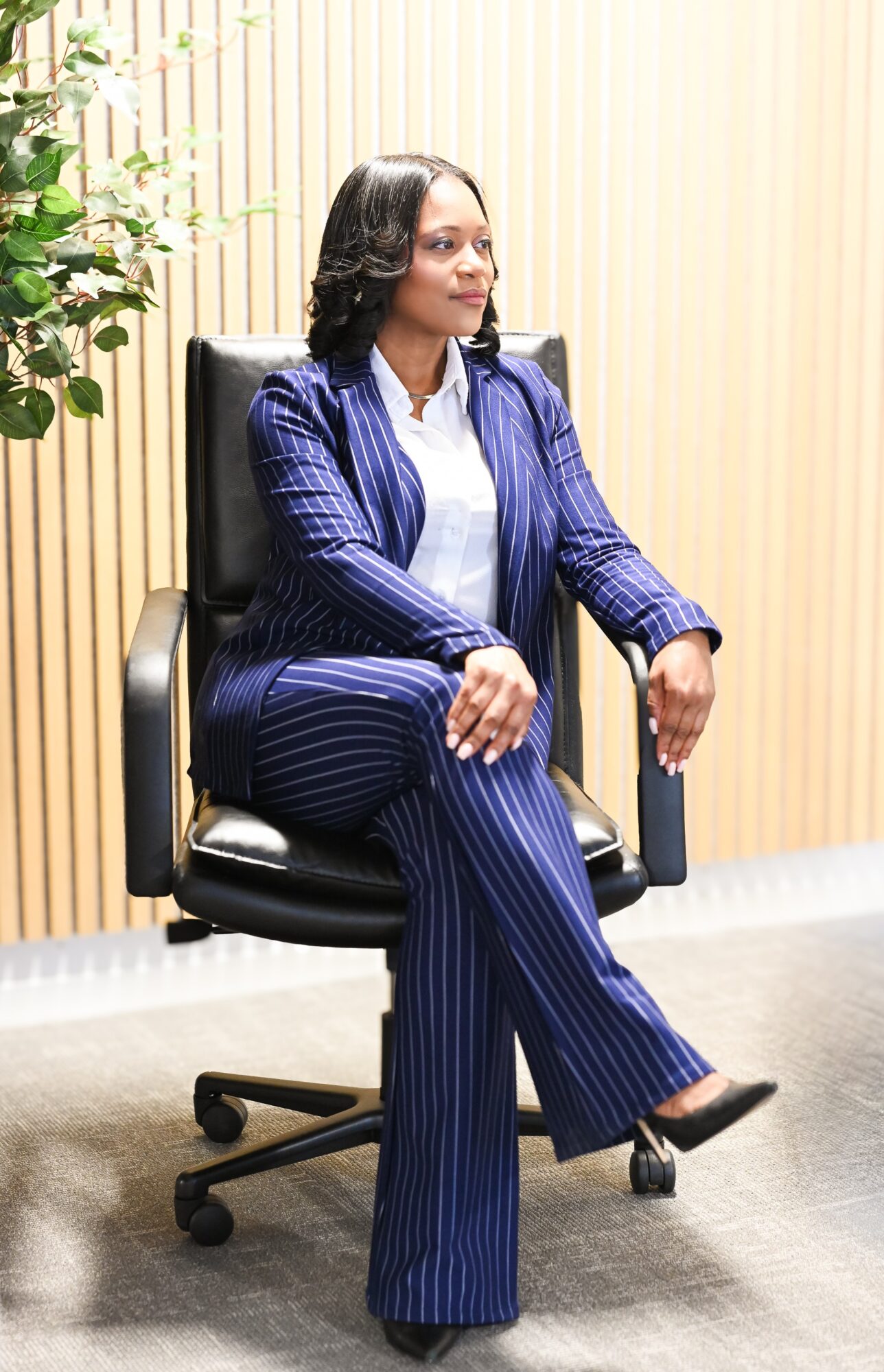 Woman sitting in a black office chair with crossed legs, wearing a blue pinstripe suit, in a room with light-colored walls and a plant.
