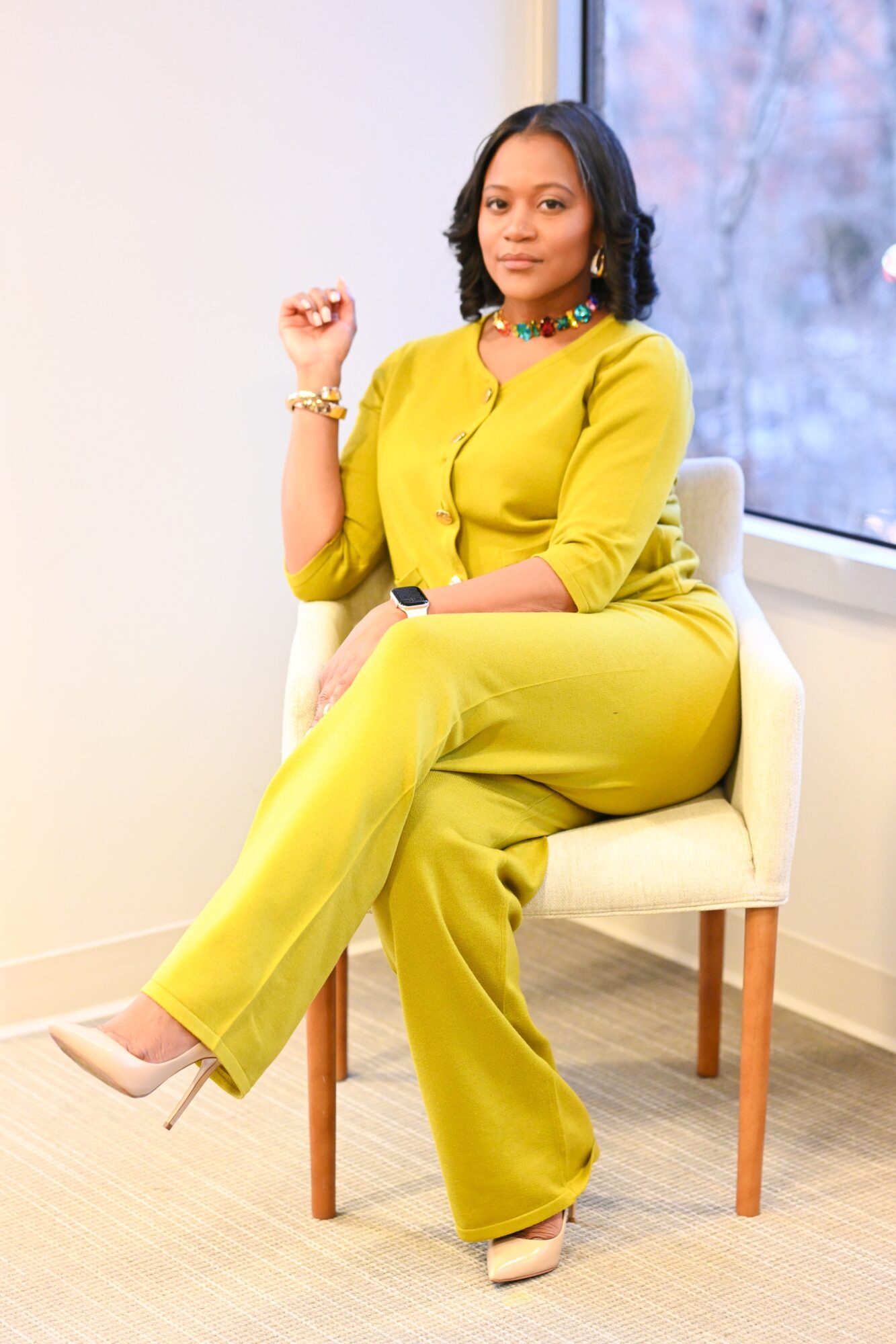 Woman in yellow outfit sitting on a chair indoors near a window, with a neutral background.
