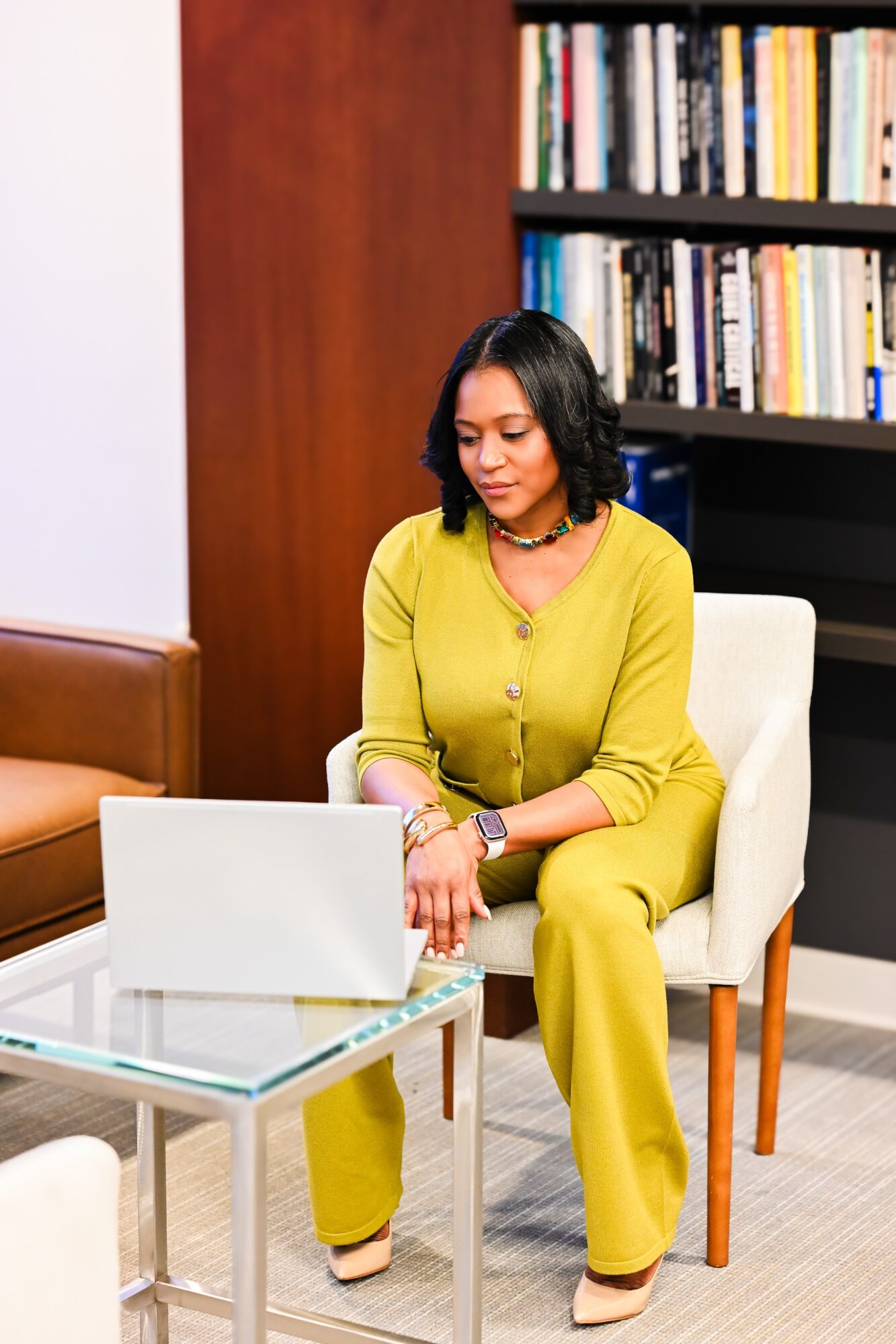 Woman in yellow outfit sitting in a chair, working on a laptop in a room with bookshelves.