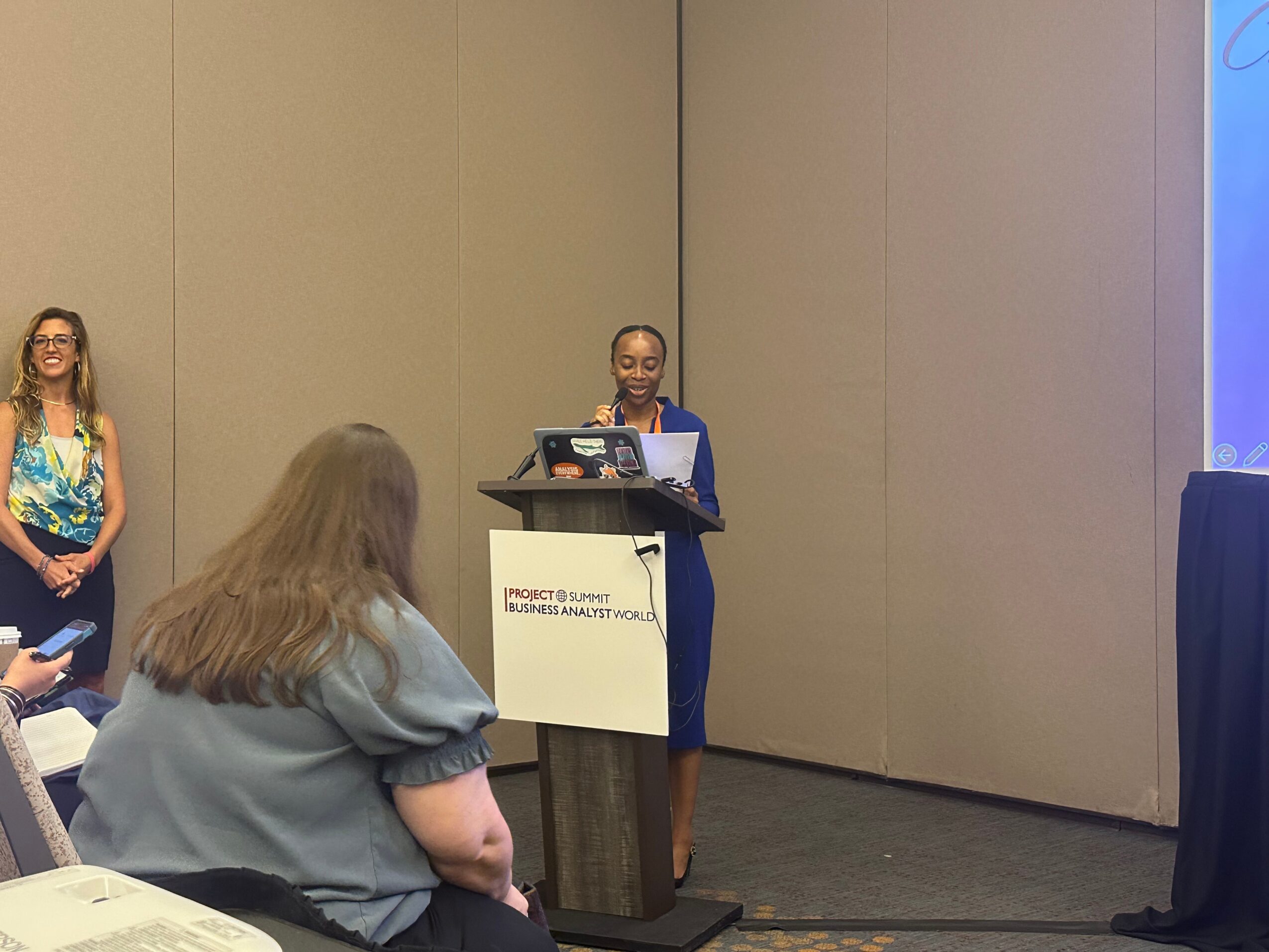 A woman stands at a podium giving a presentation in a conference room, with a woman seated and a woman standing in the background.