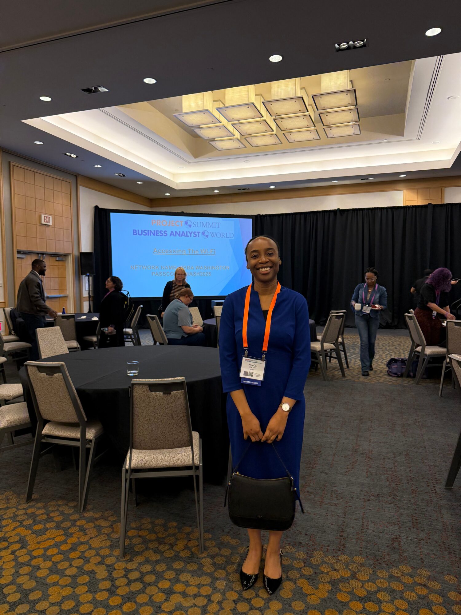 Woman in blue dress with orange lanyard standing in conference room, holding a bag, with a presentation screen behind her.